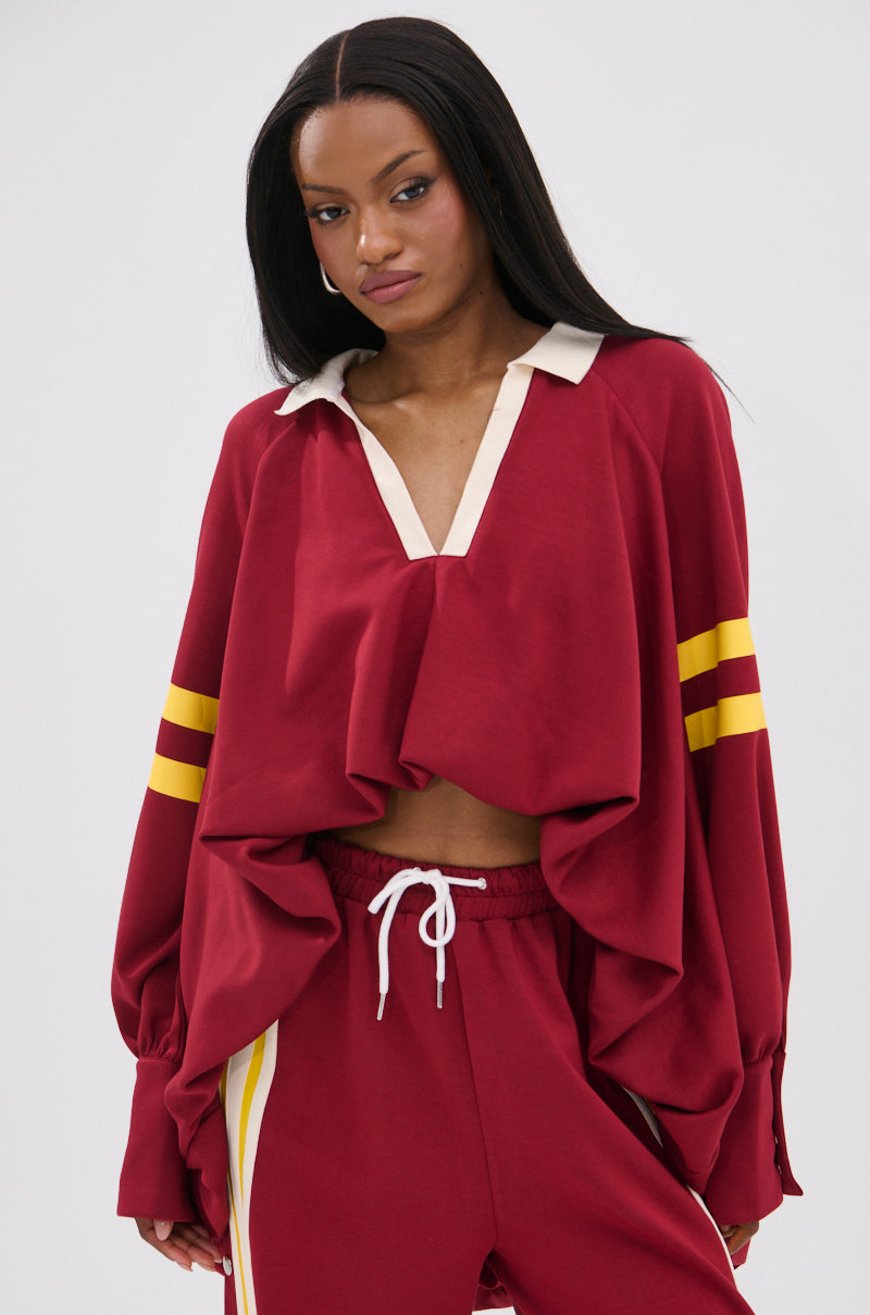 A woman wears the GAME OVER V-NECK SWEATER, a loose 90’s-inspired maroon pullover with yellow striped sleeves, cream collar, and wide sleeves, paired with matching maroon pants featuring varsity white and yellow side stripes.