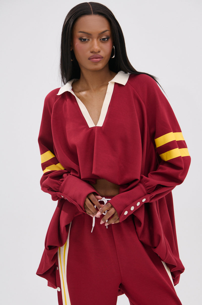 A woman in the GAME OVER V-NECK SWEATER—loose-fitting with maroon and yellow stripes—stands against a plain background, varsity-inspired. She has straight, long hair, a neutral expression, and her hands clasped in front.