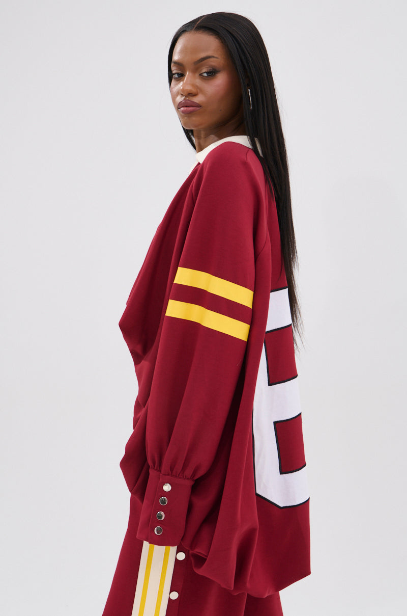 A woman with long straight hair wears the loose-fitting, 90’s-inspired maroon GAME OVER V-NECK SWEATER, featuring yellow sleeve stripes and bold numbers on the back, paired with matching pants for varsity vibes against a plain background.