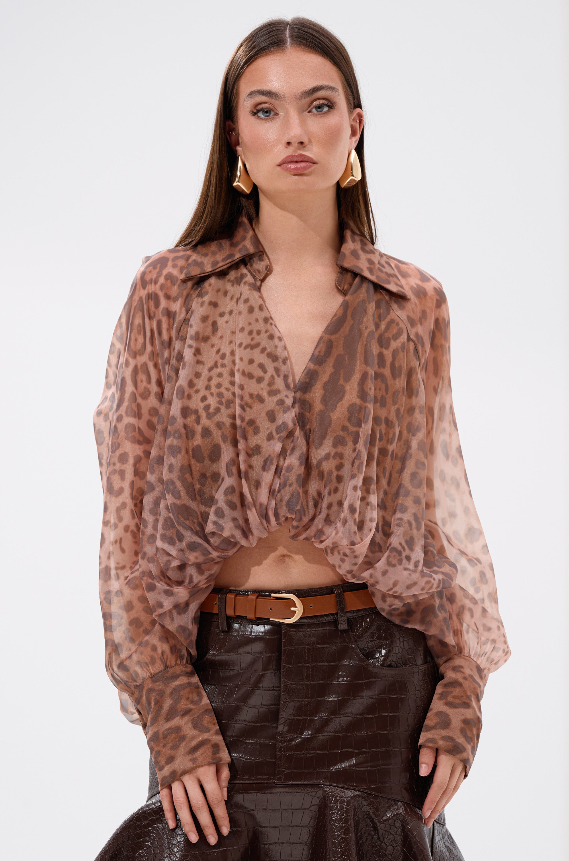 A woman wears the WILD SPIRIT HIGH LOW SHEER BLOUSE in cheetah print with a brown belted textured skirt, standing against a plain white background.