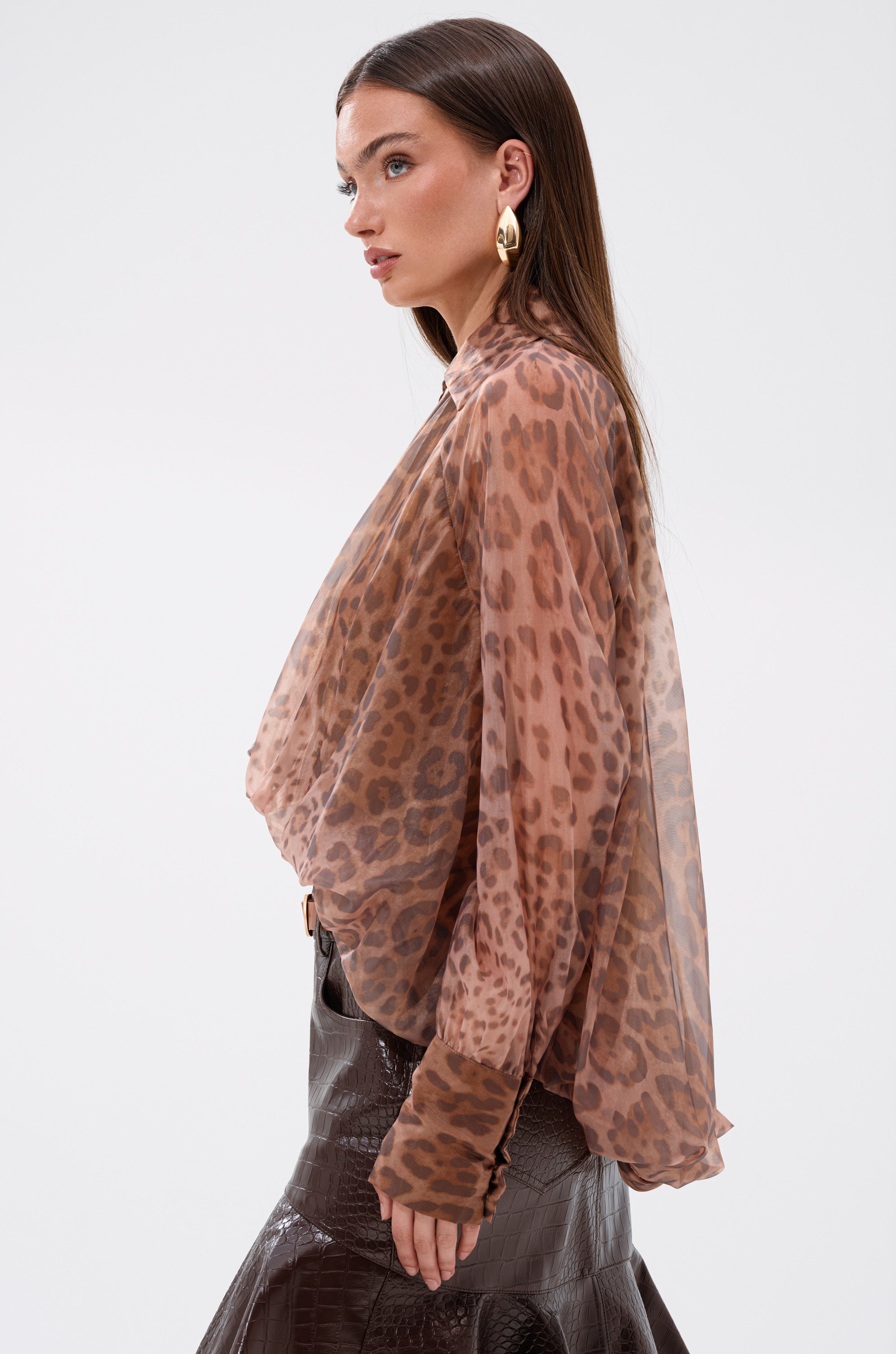 A woman with long brown hair models the WILD SPIRIT HIGH LOW SHEER BLOUSE in a cheetah print and a shiny textured dark brown skirt, standing in profile against a plain white background.
