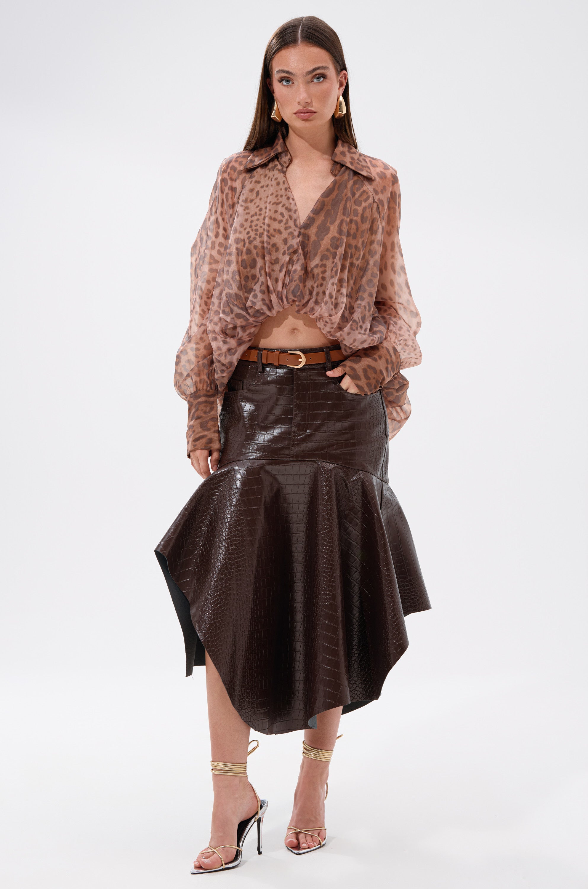 A woman stands against a plain white background wearing the AKIRA Label WILD SPIRIT HIGH LOW SHEER BLOUSE, a brown asymmetrical faux leather skirt, a gold belt, and strappy high-heeled sandals.