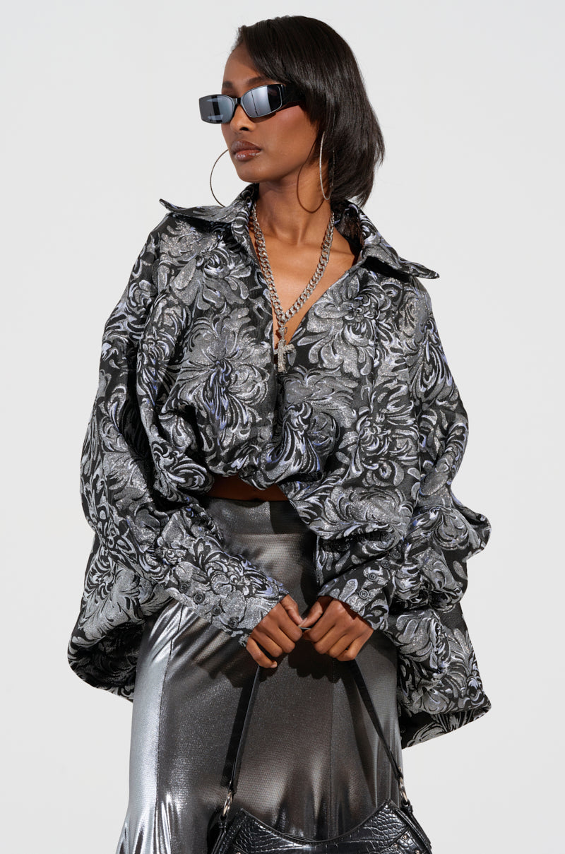 A person wearing oversized sunglasses pairs the WHO WE ARE BLOUSE, with its high-low silhouette, beneath a floral-patterned metallic jacket. They style it with a shiny silver skirt, accessorize with a textured handbag, large hoop earrings, and a chain necklace while standing against a plain background.