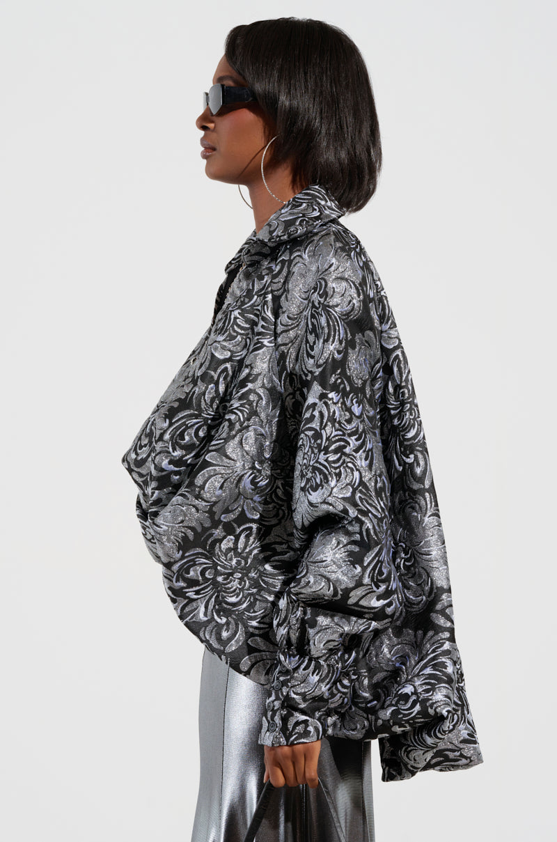 A person wearing oversized sunglasses and large hoop earrings poses in profile, showcasing a metallic brocade-patterned oversized jacket reminiscent of the WHO WE ARE BLOUSE, paired with shiny silver pants against a simple backdrop.