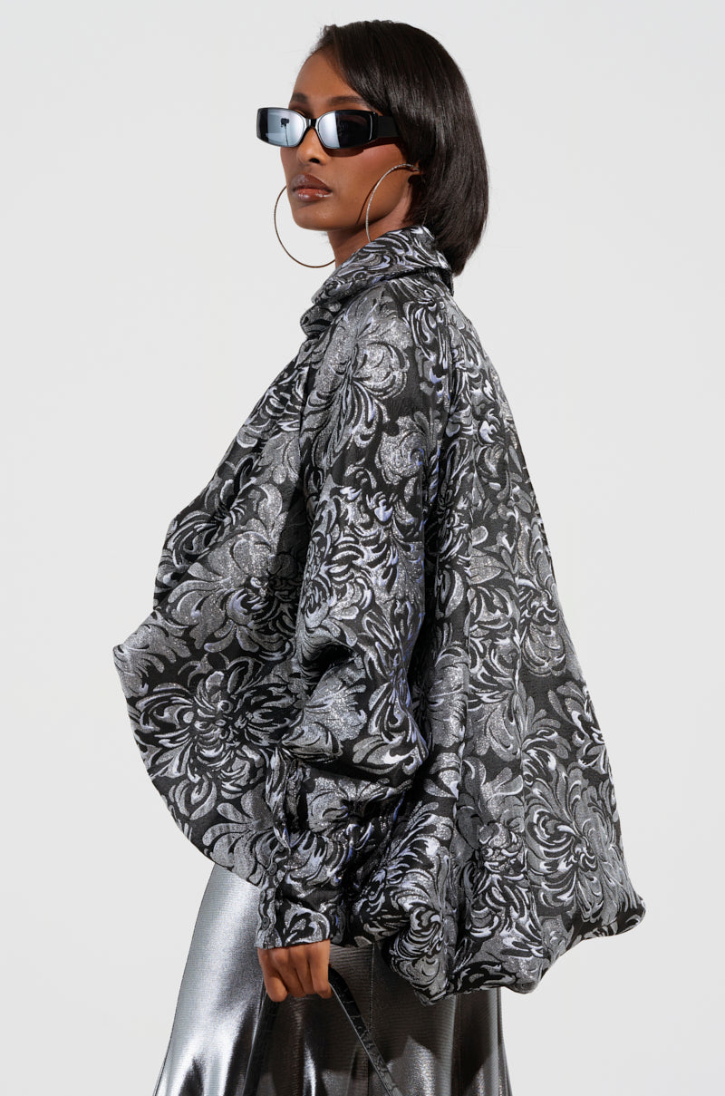 A person wearing oversized sunglasses, gold hoop earrings, and a metallic brocade pattern jacket over the WHO WE ARE BLOUSE from AKIRA Label is striking a pose. The ensemble is completed with a metallic skirt, exuding confidence and style against a plain background.