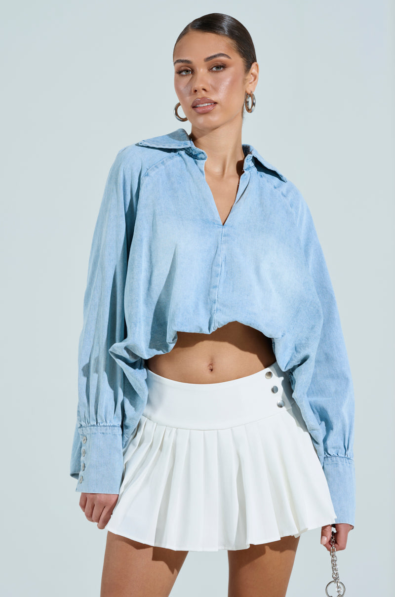 Someone wears an AMELIA DENIM HI LOW BLOUSE in light blue, paired with a white pleated mini skirt. They accessorize with hoop earrings and have their hair styled back. The solid light background accentuates the blouse's high-low silhouette.