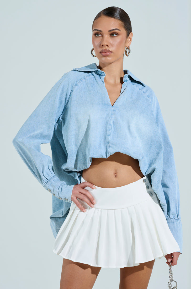 A woman poses against a neutral background in the AMELIA Denim Hi Low Blouse, a light blue cropped cotton blend, paired with a white pleated skirt. Her hand on her hip and a sideways gaze effortlessly captures modern elegance.