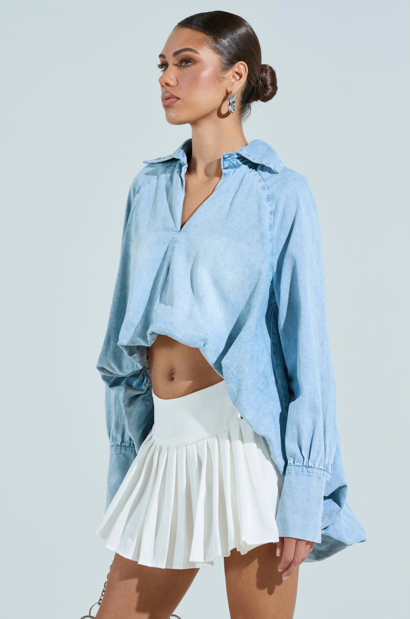 A woman stands against a light background wearing the AMELIA DENIM HI LOW BLOUSE, an oversized light blue cotton blend shirt. She pairs it with a white pleated skirt and large silver star-shaped earrings. Her hair is tied back as she gazes to the side confidently.