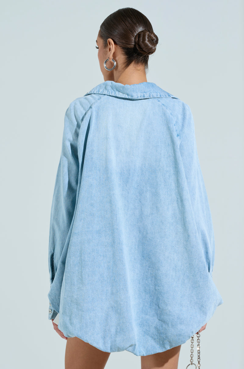 A person with smooth hair tied in a bun wears the AMELIA DENIM HI LOW BLOUSE. They face away, highlighting the cotton blend denim's draped back and chic high-low silhouette against a solid blue backdrop.