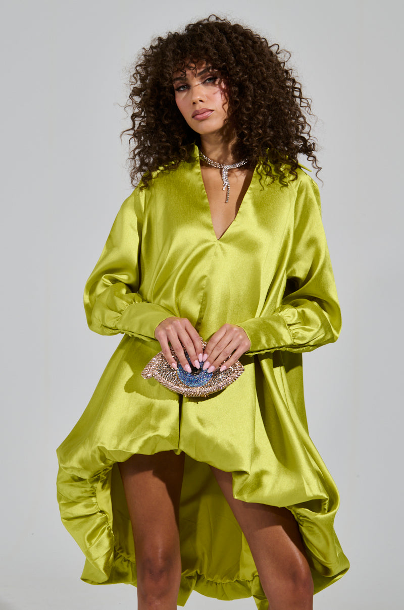 A woman with curly hair wears an Elouise Hi Low Dress in green, the flowing silk creating a subtle bubble silhouette. She holds a small embellished clutch and wears a silver necklace, all set against a plain light gray background.