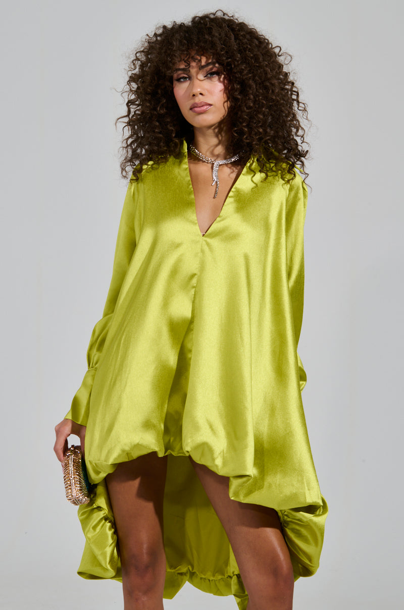 A woman with curly hair is wearing the ELOUISE HI LOW DRESS IN GREEN, which features a bubble silhouette. She holds a small clutch and poses against a plain background, accessorized with a statement necklace.