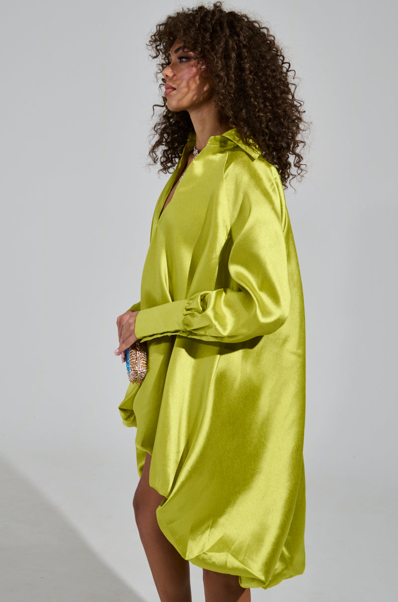 A person with curly hair stands in profile, donning the vibrant ELOUISE HI LOW DRESS IN GREEN, crafted from shiny satin with long sleeves. They clutch a small colorful bag against the plain gray backdrop.