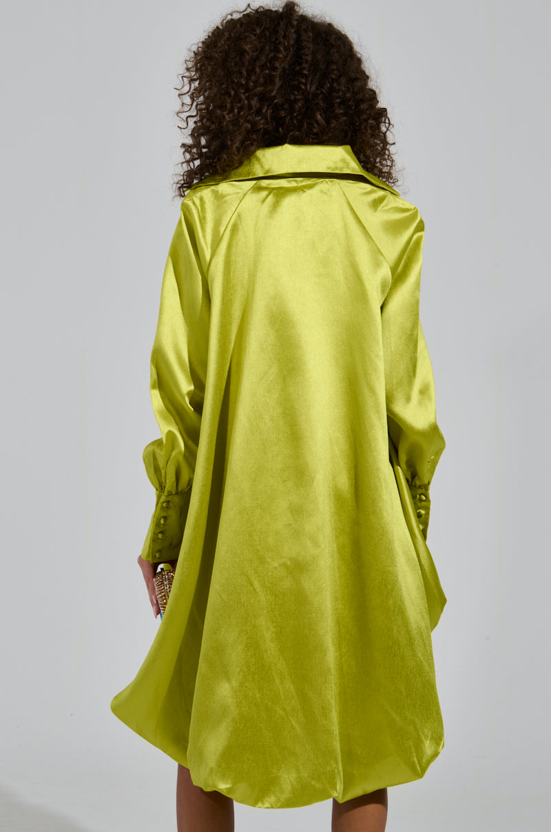Back view of a person with curly hair wearing the ELOUISE HI LOW DRESS IN GREEN, showcasing its bubble silhouette. The long sleeves are adorned with decorative buttons, and the ensemble is completed with an embellished clutch in hand.