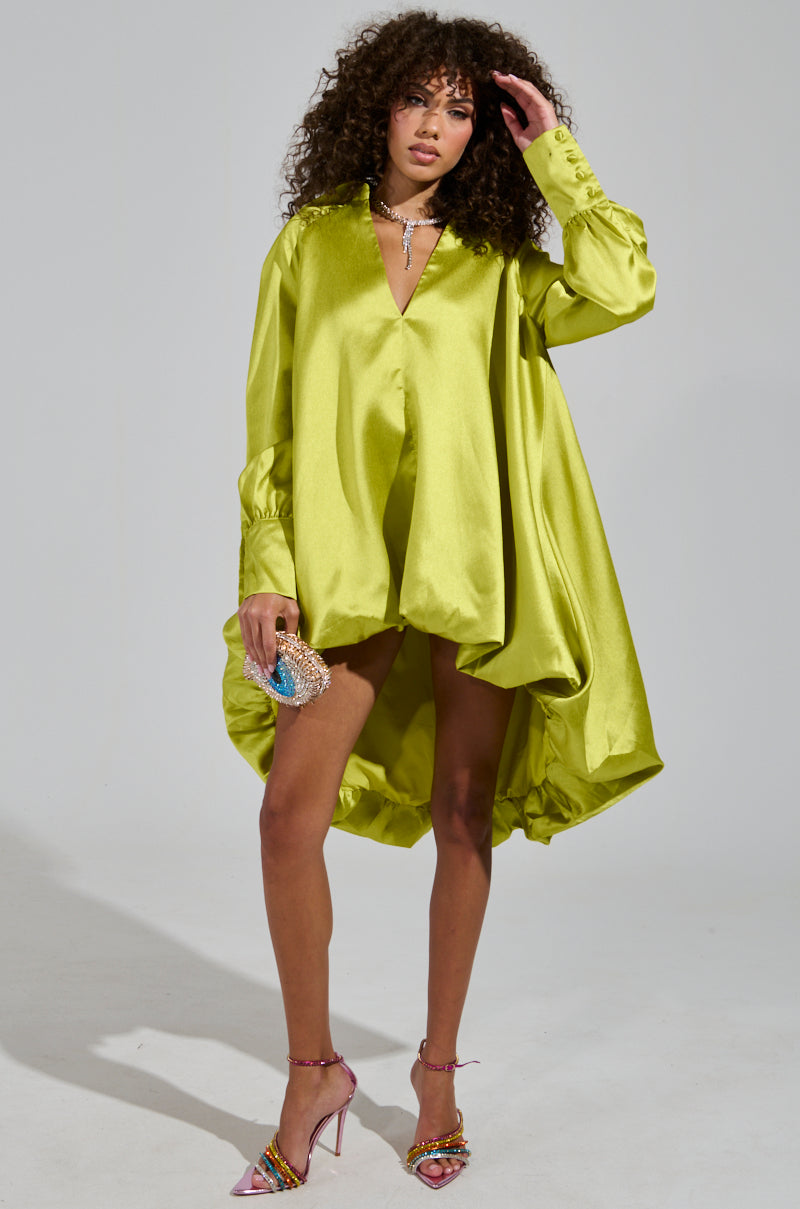 A woman with curly hair poses in a loose-fitting, green ELOUISE HI LOW DRESS IN GREEN. She is holding a small, embellished purse and wearing pink high heels. The dress's bubble silhouette contrasts beautifully with the simple, gray gradient background.