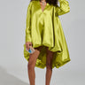 A woman with curly hair poses in a loose-fitting, green ELOUISE HI LOW DRESS IN GREEN. She is holding a small, embellished purse and wearing pink high heels. The dress's bubble silhouette contrasts beautifully with the simple, gray gradient background.