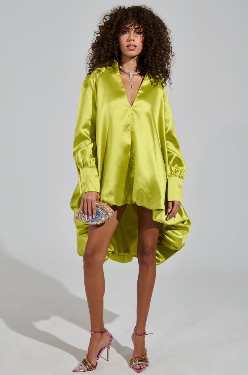 A person with curly hair stands against a plain background, wearing an ELOUISE HI LOW DRESS IN GREEN featuring a bubble silhouette. They hold a glittery clutch and wear colorful high-heeled sandals.