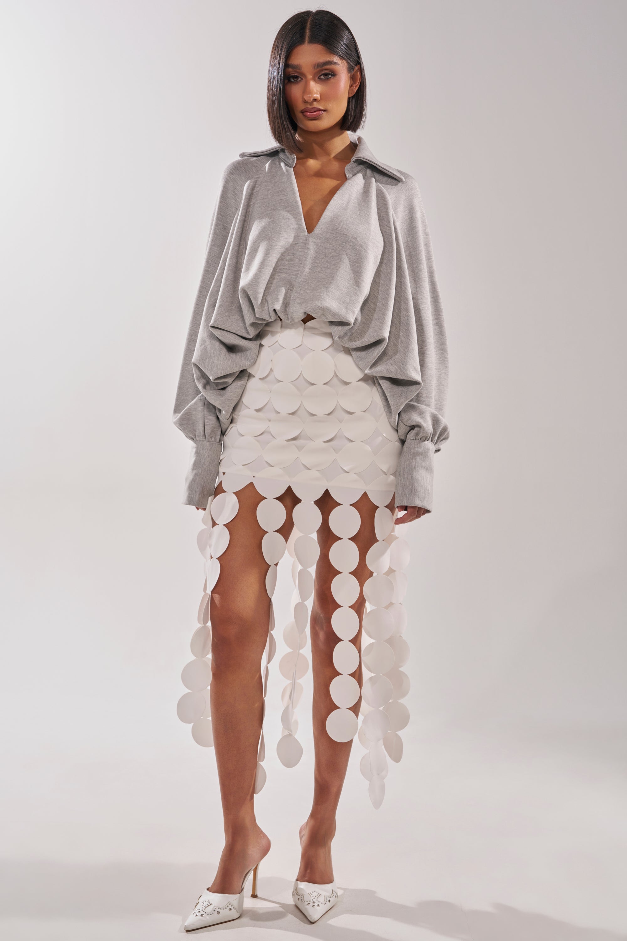 A woman models the NEW RULES HIGH LOW BLOUSE IN GREY, a loose, oversized-sleeved top paired with a white skirt featuring large circular discs and white pointed-toe heels, standing against a plain light background. 5