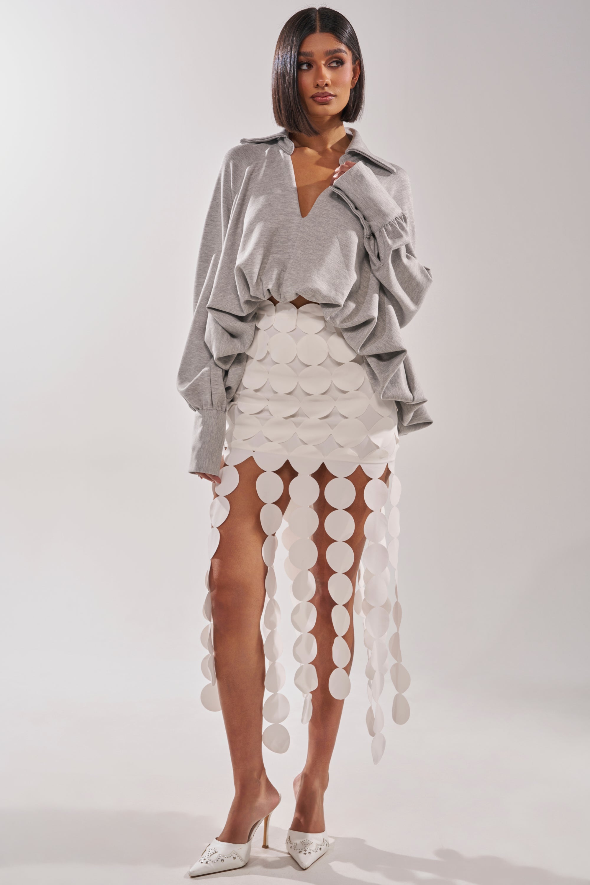 A woman wears the NEW RULES HIGH LOW BLOUSE IN GREY, styled with a distinctive white skirt featuring circular cutouts and white circles on vertical strips, paired with white pointed-toe heels against a plain light background. 6