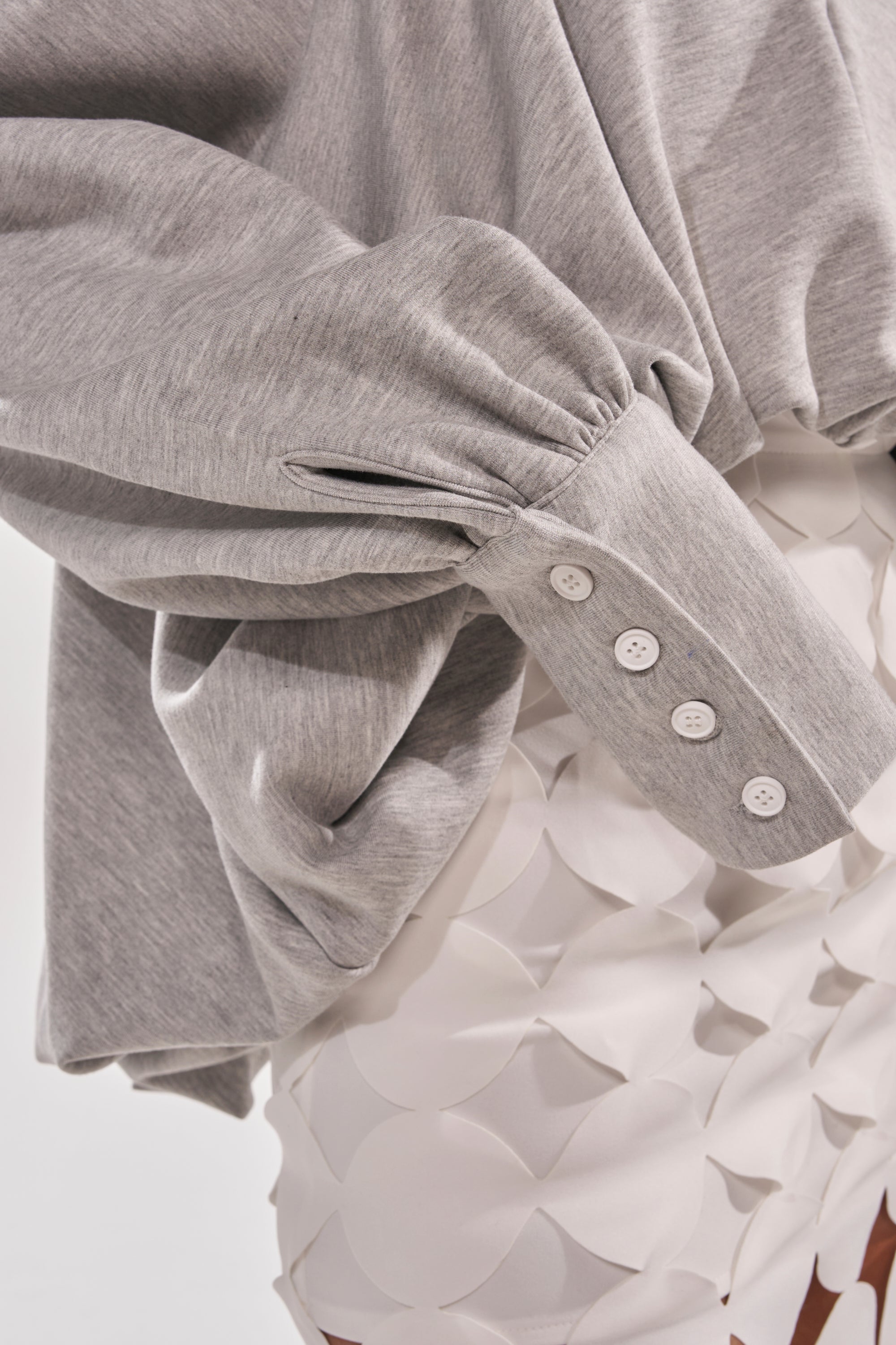 A close-up of a person wearing the NEW RULES HIGH LOW BLOUSE IN GREY with buttoned cuffs and an asymmetrical hem, paired with a white skirt featuring layered circular fabric details. The background is plain and light-colored. 7