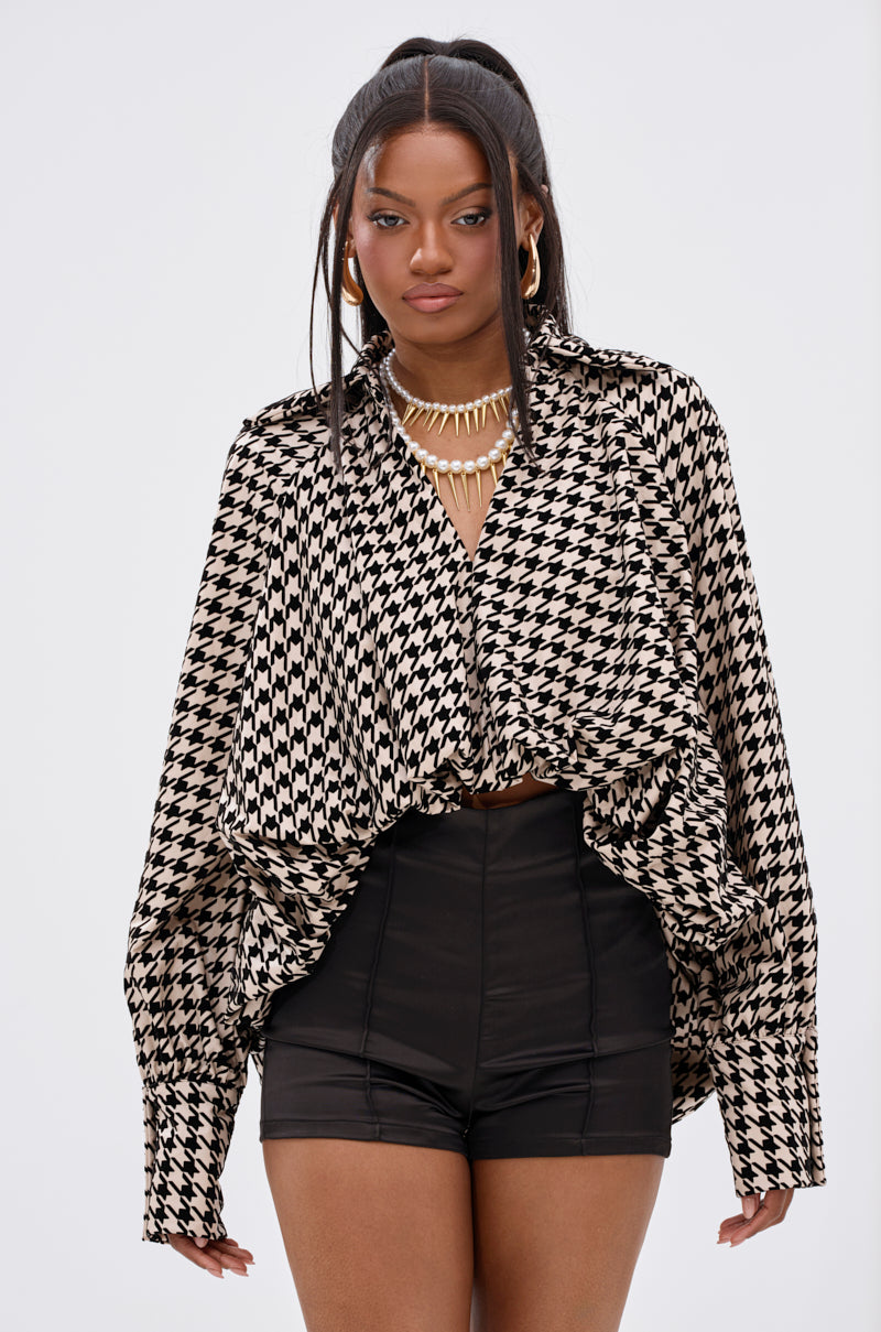 A confident woman styles the I KNOW WHAT I WANT HOUNDSTOOTH BLOUSE TOP, featuring a loose fit, asymmetrical high-low hem, and wide long sleeves, with black high-waisted shorts, statement jewelry, and her dark hair in a ponytail.