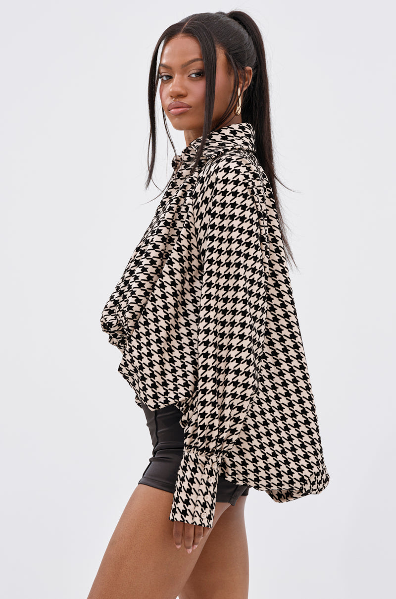 A woman with long dark hair in a high ponytail wears the I KNOW WHAT I WANT HOUNDSTOOTH BLOUSE TOP and black shorts, standing against a plain white background.