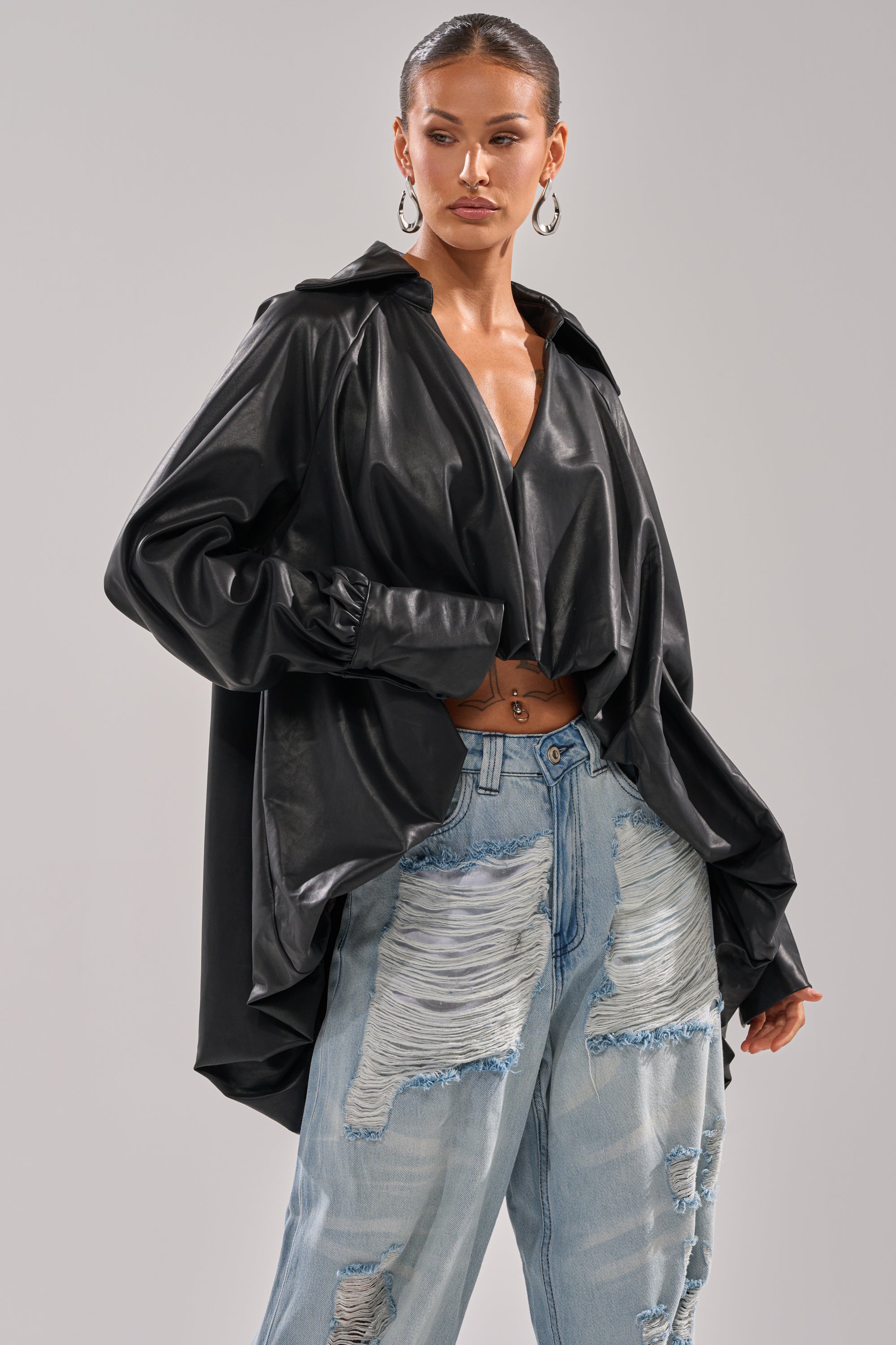A woman models the NEW ATTITUDE FAUX LEATHER HI-LOW BLOUSE with voluminous sleeves, styled with light blue ripped jeans, slicked-back hair, hoop earrings, and a navel piercing.