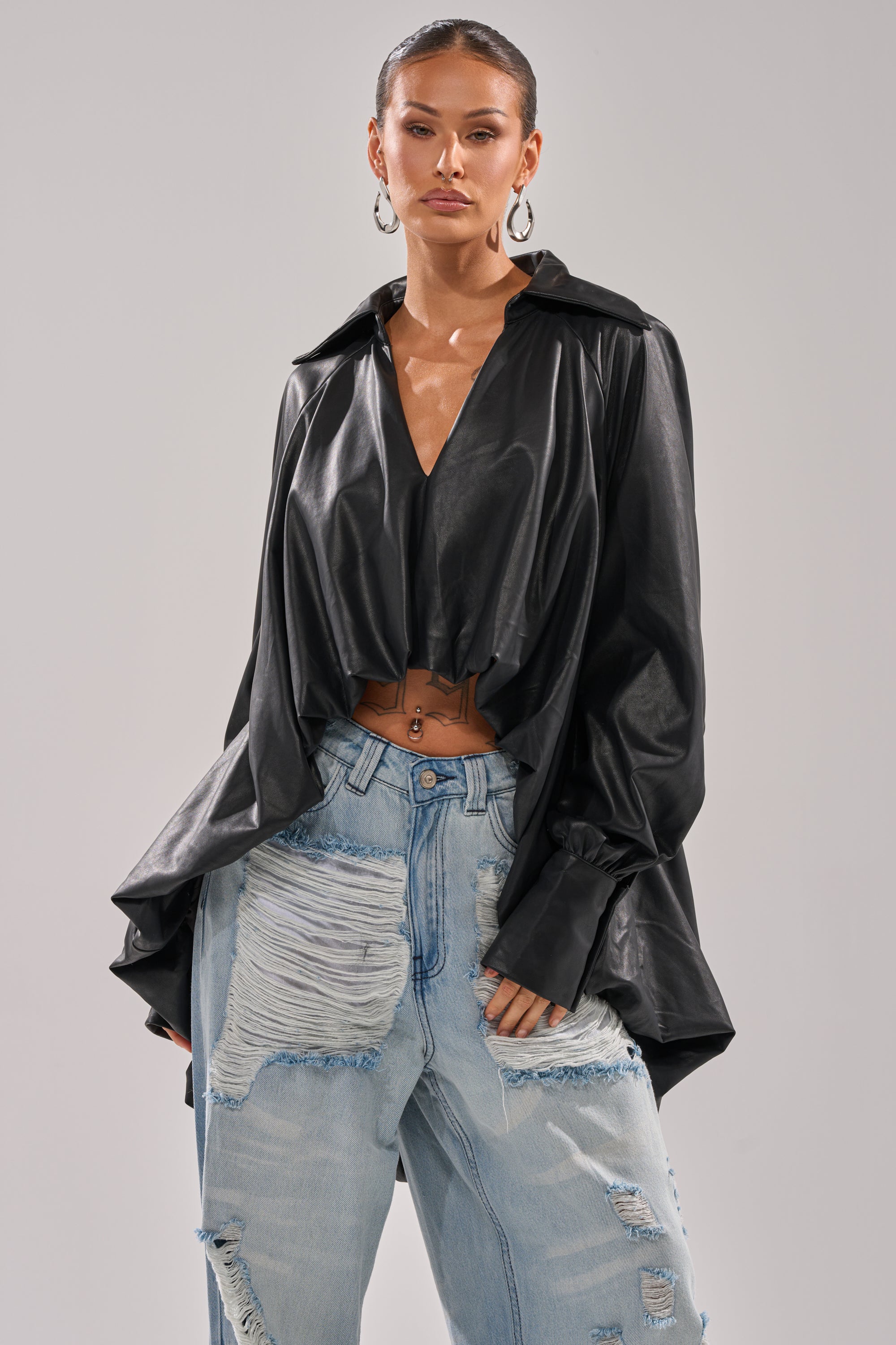 Wearing the NEW ATTITUDE FAUX LEATHER HI-LOW BLOUSE in black with a deep V-neck, paired with light blue distressed high-waisted jeans and large hoop earrings, a person stands against a plain background.