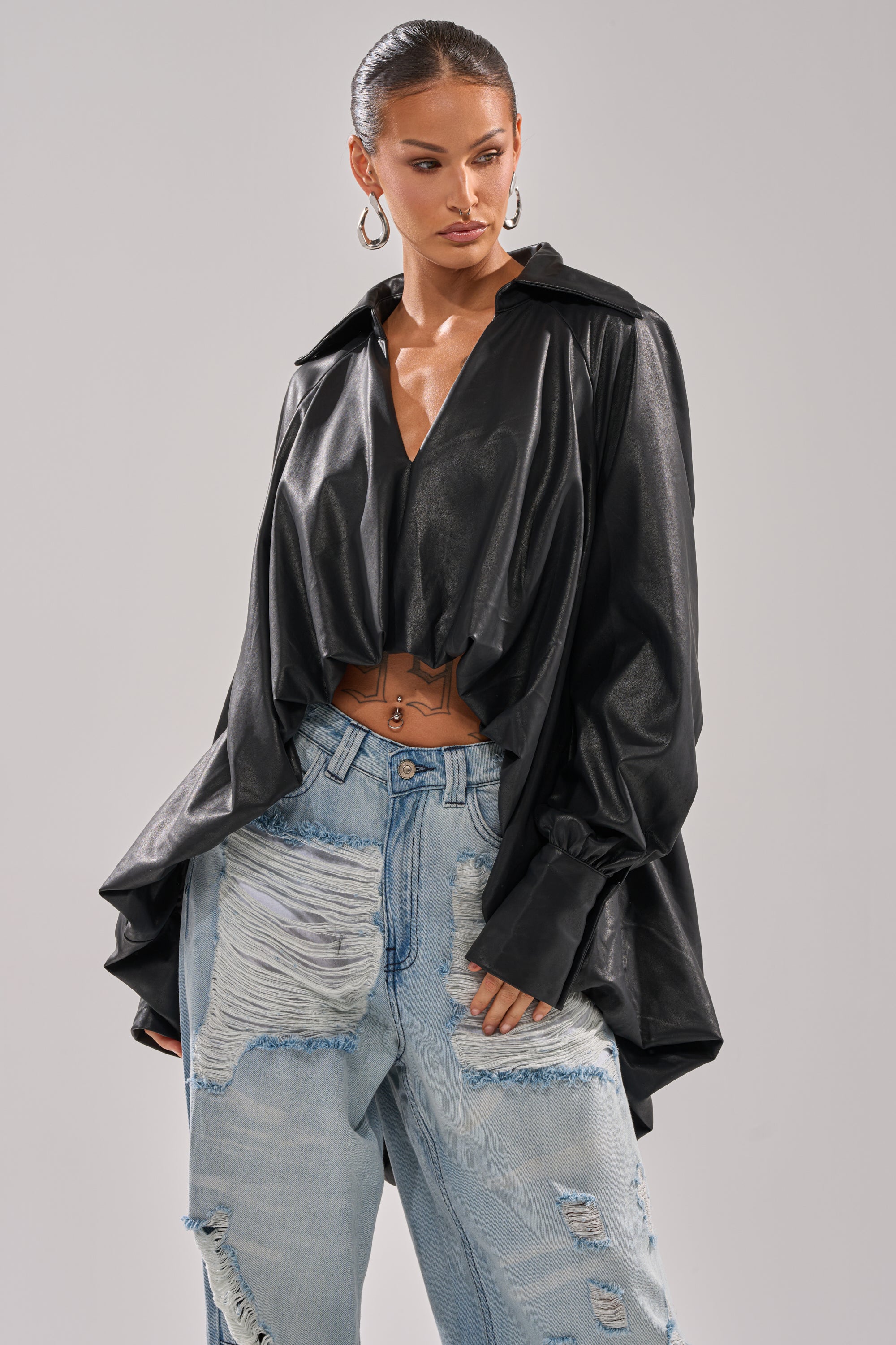 A woman models the NEW ATTITUDE FAUX LEATHER HI-LOW BLOUSE, a loose black top with wide sleeves, paired with light blue ripped jeans and hoop earrings, striking an effortlessly chic pose against a plain grey background.