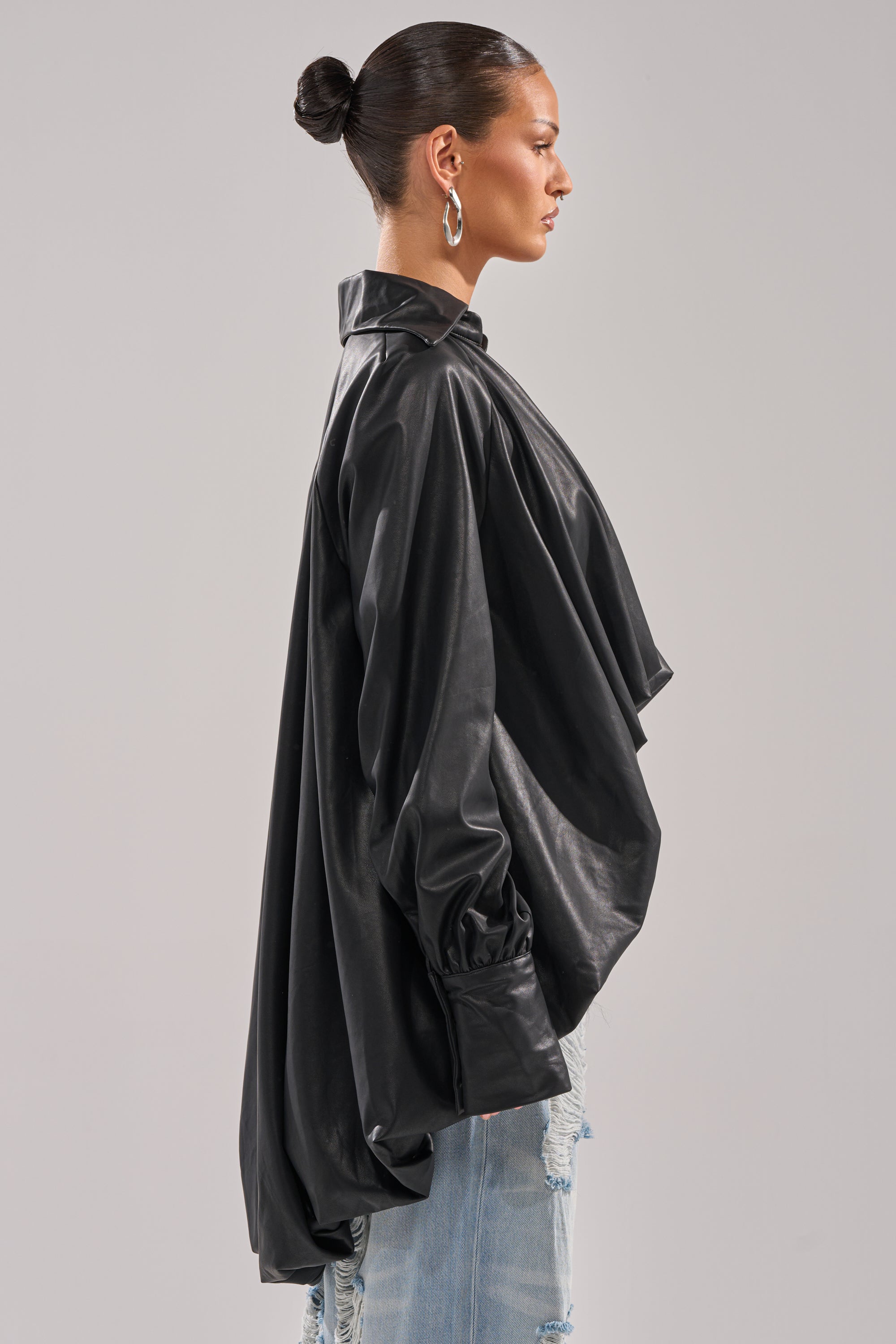 A woman with her hair in a sleek bun models the NEW ATTITUDE FAUX LEATHER HI-LOW BLOUSE, a loose-fitting black top with oversized sleeves, styled with light-wash distressed jeans and shown in profile against a plain background.