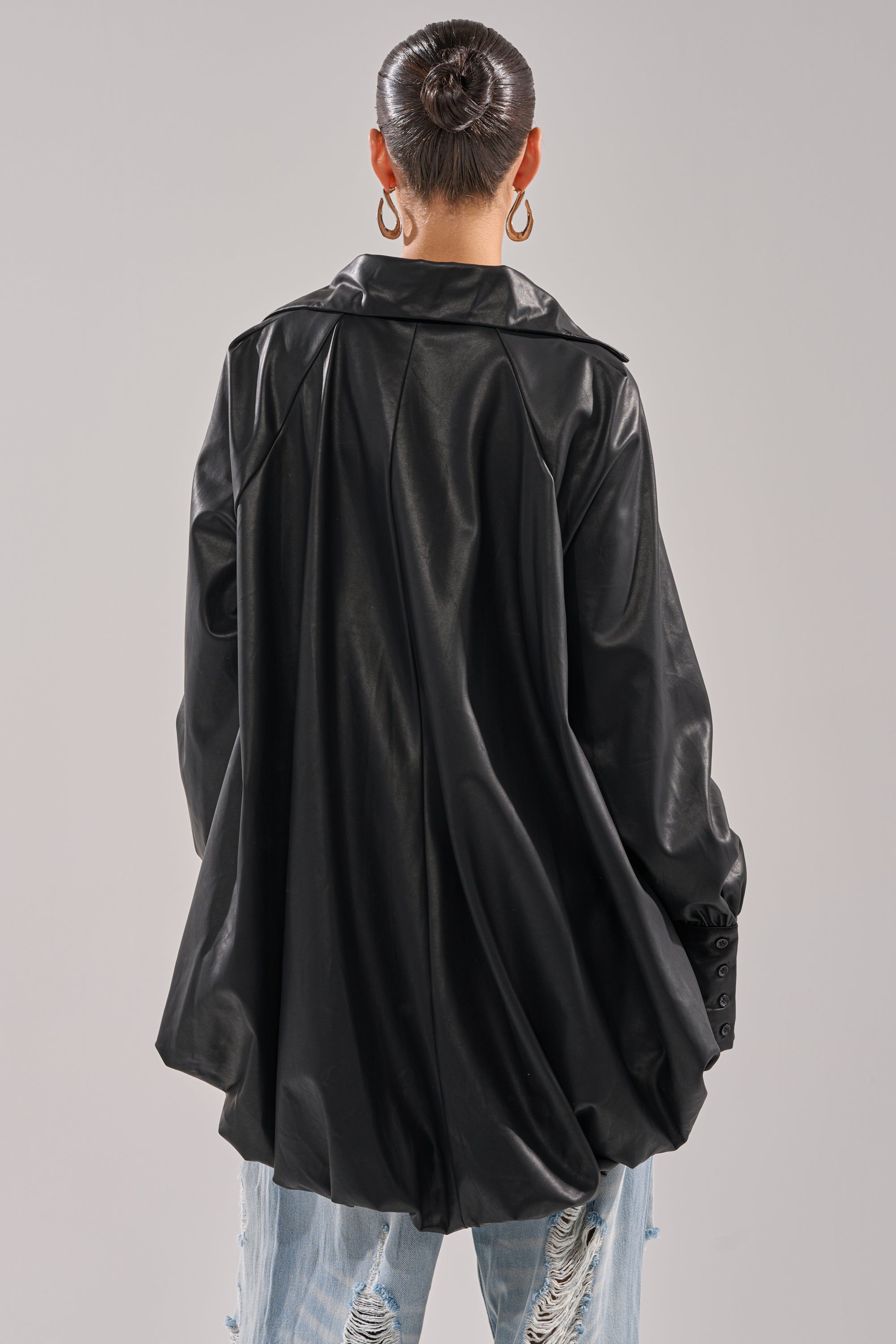 A person with their hair in a sleek bun models the NEW ATTITUDE FAUX LEATHER HI-LOW BLOUSE in black, paired with light blue distressed jeans, facing away from the camera against a plain light background.