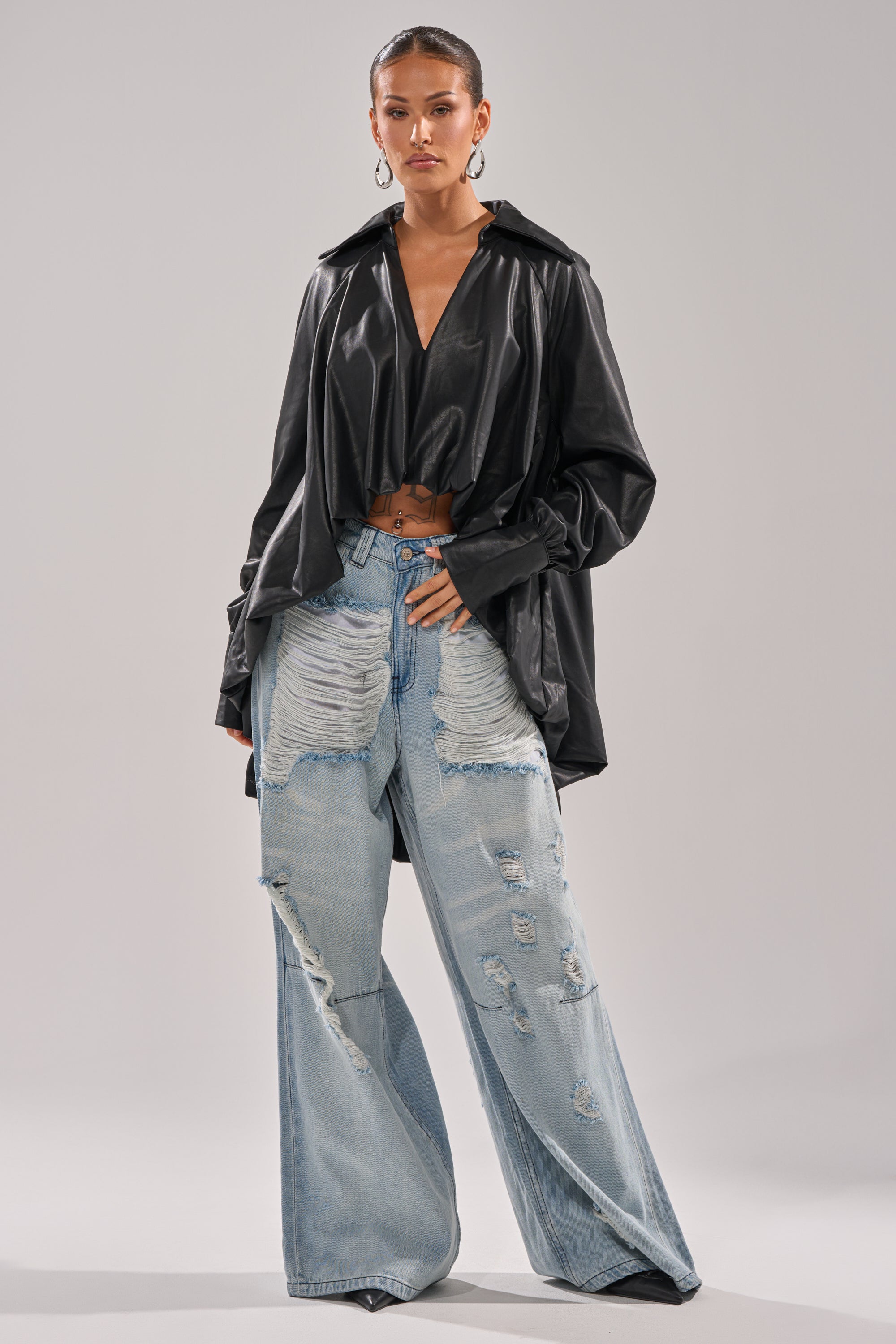 A woman models the NEW ATTITUDE FAUX LEATHER HI-LOW BLOUSE in black with wide sleeves, styled with light blue, high-waisted distressed wide-leg jeans and hoop earrings, standing against a plain background.