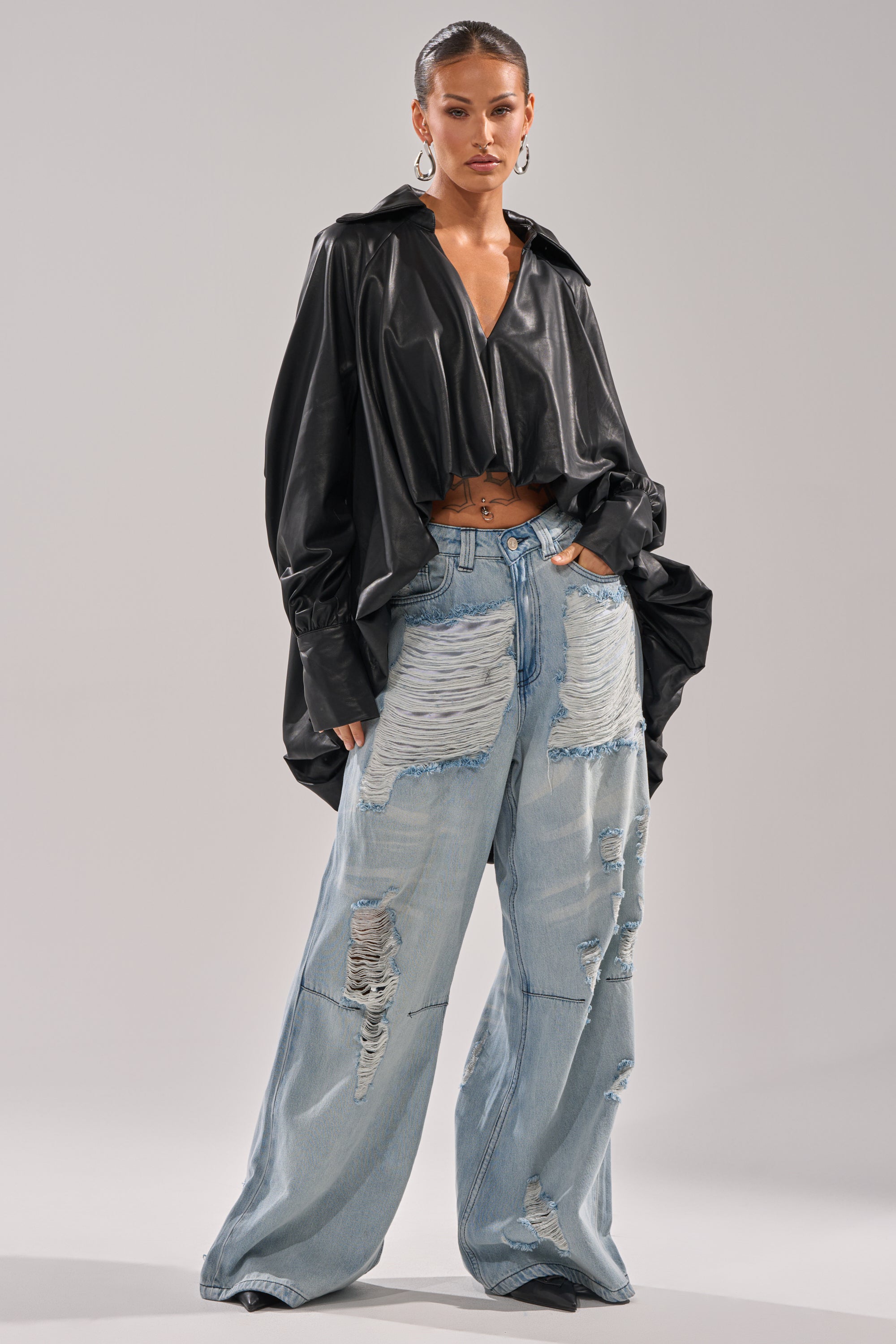 A woman wears the NEW ATTITUDE FAUX LEATHER HI-LOW BLOUSE with wide sleeves and pairs it with high-waisted, light blue distressed wide-leg jeans, standing confidently against a plain light background.