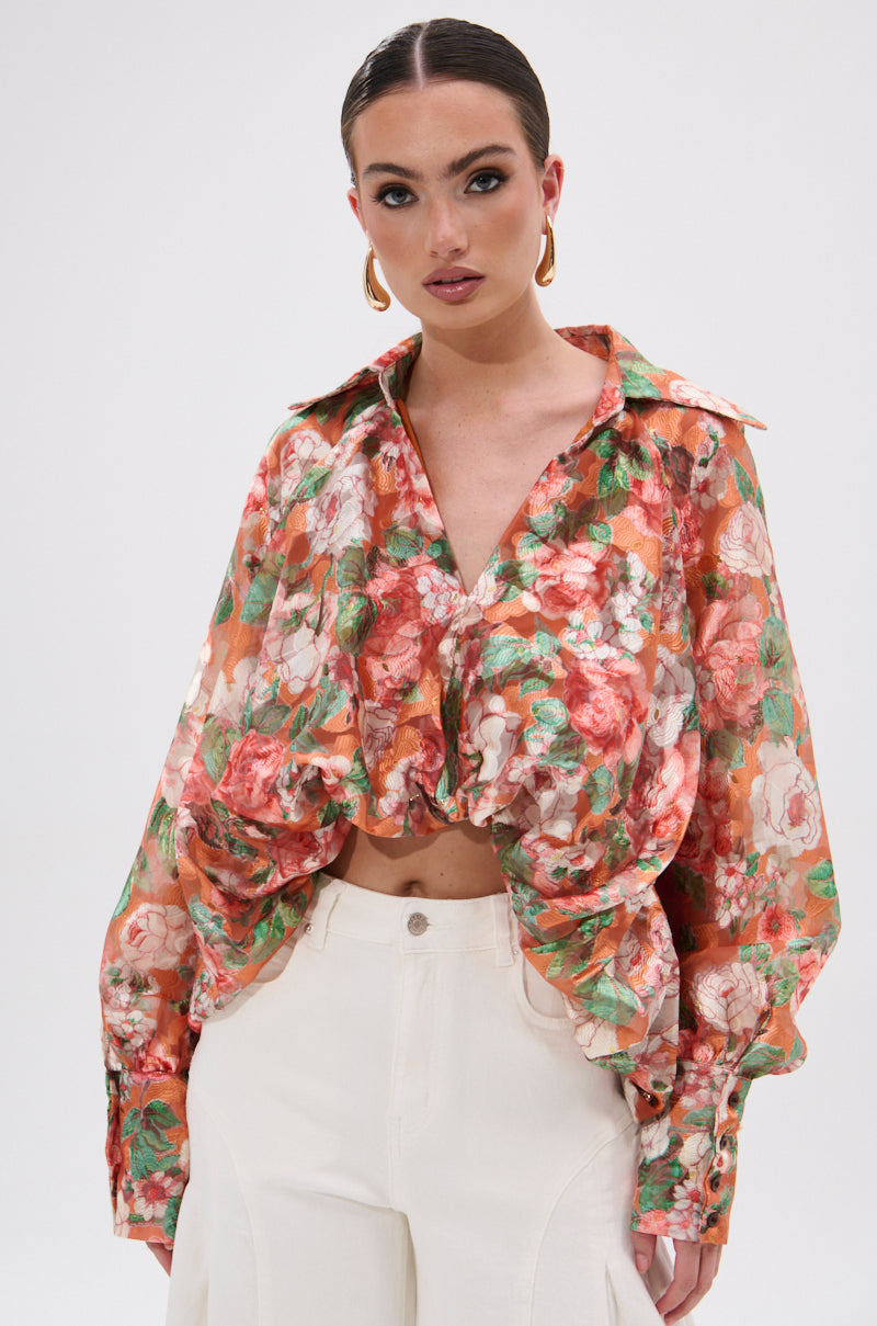 A woman wears the SUNSHINE FLORAL HIGH LOW BLOUSE, a sheer floral top with long balloon sleeves in red, green, and orange, paired with high-waisted white pants. She completes her look with gold hoop earrings and slicked-back dark hair against a plain white background.