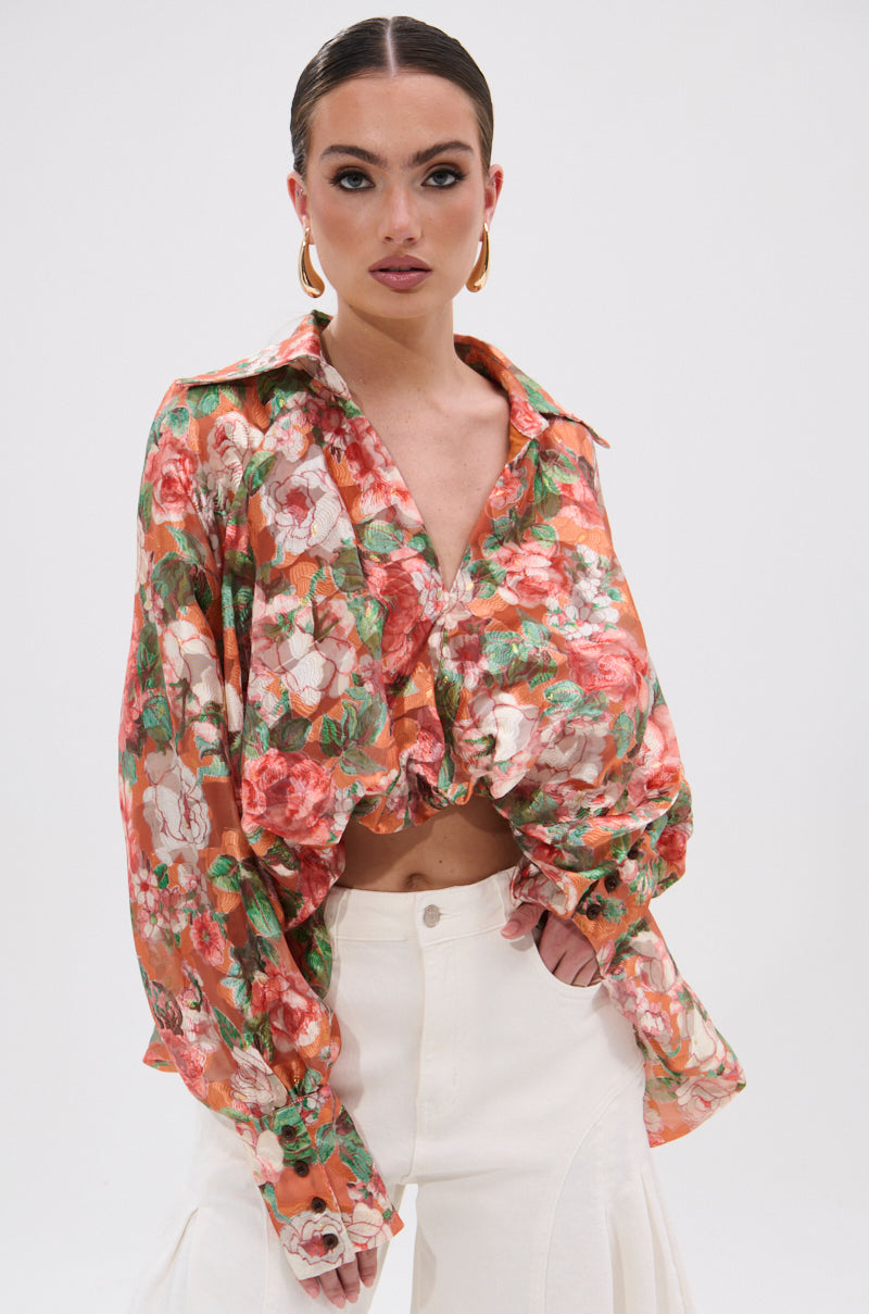 A woman models the SUNSHINE FLORAL HIGH LOW BLOUSE with balloon sleeves, paired with white high-waisted pants. She poses against a plain white background, her hair slicked back and accessorized with gold hoop earrings.