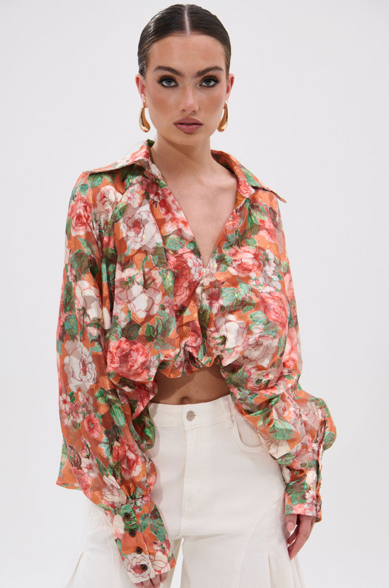 A woman with slicked-back hair wears gold hoop earrings and the SUNSHINE FLORAL HIGH LOW BLOUSE with balloon sleeves and a ruched front, paired with high-waisted white pants, standing against a plain white background.