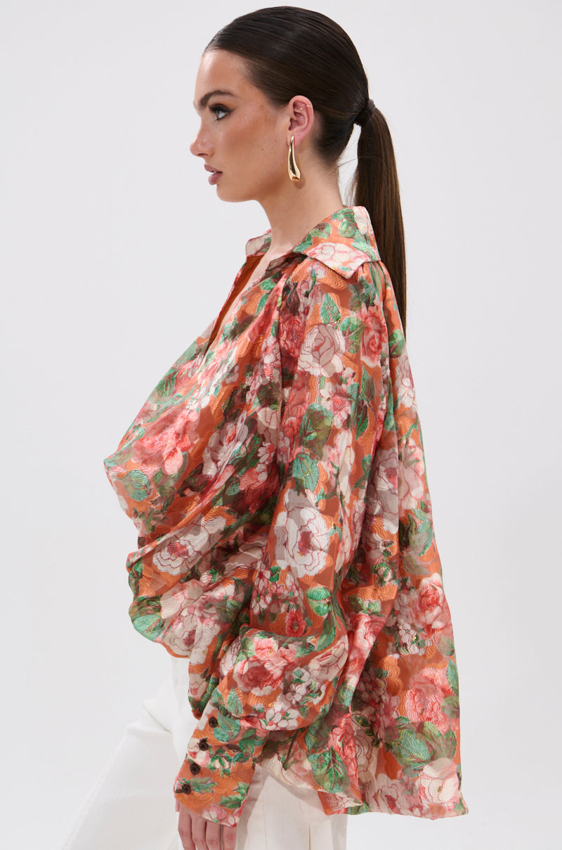 A woman in profile models the SUNSHINE FLORAL HIGH LOW BLOUSE, a sheer, long-sleeved top with pink, red, and green floral patterns and balloon sleeves. She pairs it with white pants against a plain white background.