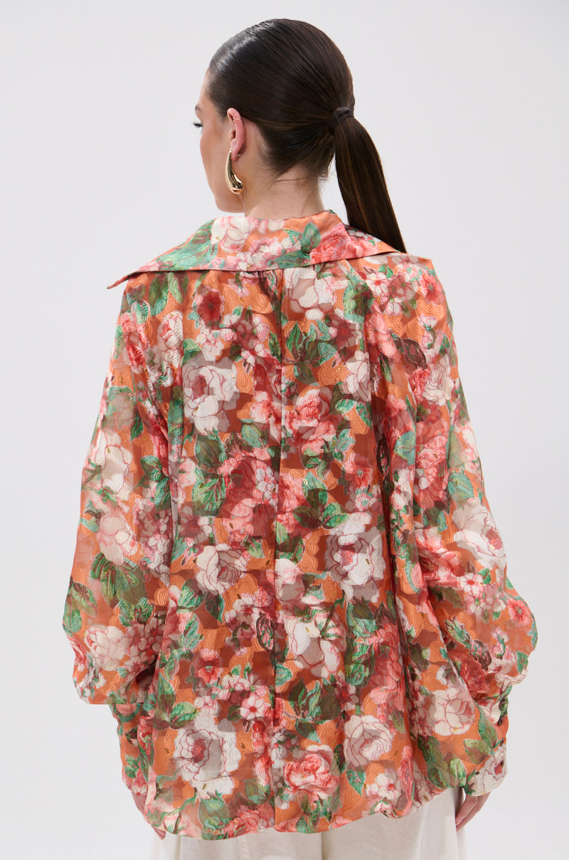 A woman with dark hair in a low ponytail is seen from behind wearing the SUNSHINE FLORAL HIGH LOW BLOUSE, a sheer top featuring balloon sleeves and a vibrant red, pink, green, and white floral print. She completes her look with gold hoop earrings.