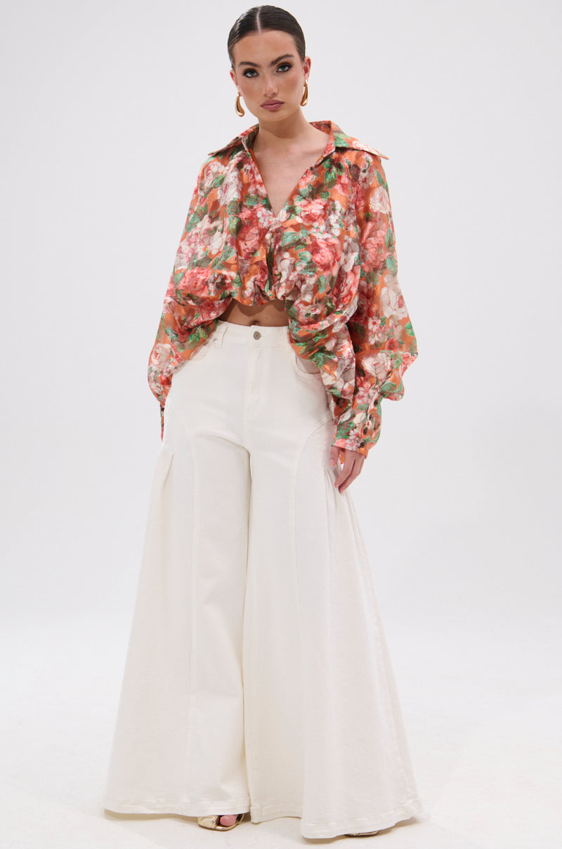 A woman models the SUNSHINE FLORAL HIGH LOW BLOUSE with balloon sleeves, a front knot, and wide-leg white pants. Her sleek hair and hoop earrings complete the confident look against a plain white backdrop.
