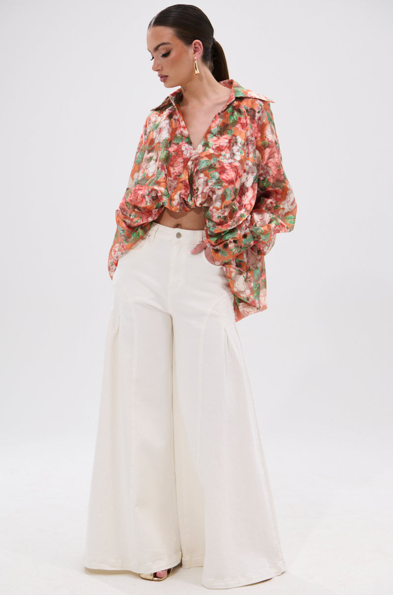 A woman models the SUNSHINE FLORAL HIGH LOW BLOUSE—a loose, balloon-sleeved top in red, green, and white—paired with high-waisted wide-leg white pants. She poses against a plain white background, looking to the side with hands in her pockets.
