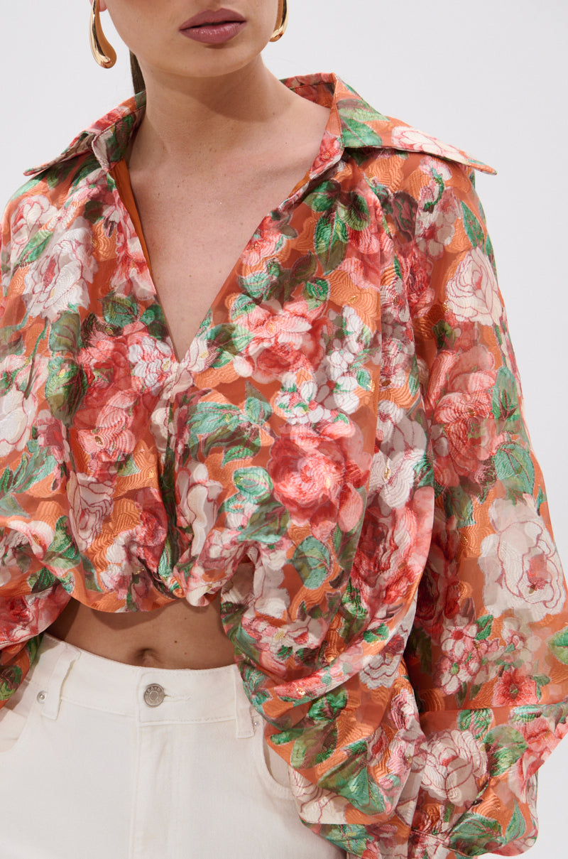 A person wears the SUNSHINE FLORAL HIGH LOW BLOUSE—a sheer, red, pink, and green floral top with balloon sleeves and a collar—paired with white high-waisted pants. The blouse’s voluminous details stand out; the person’s head is partly out of frame.