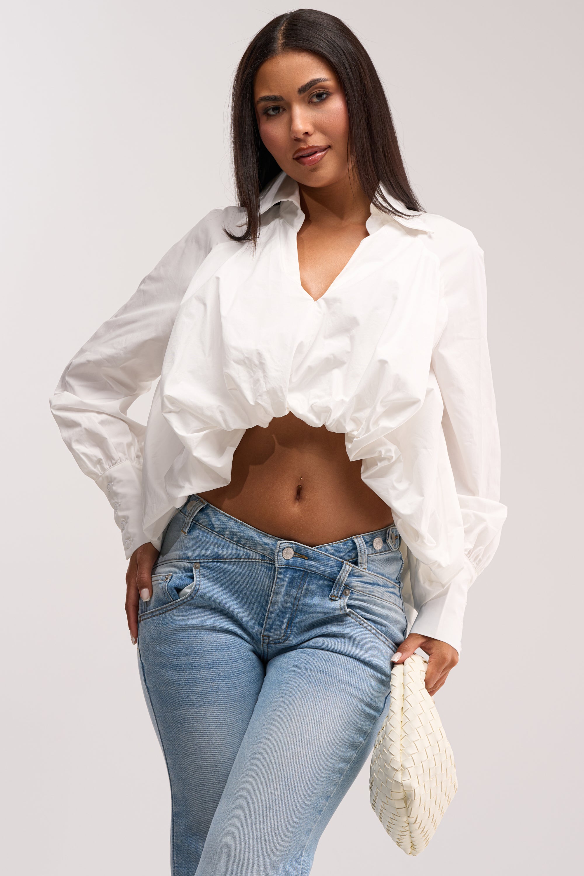 A woman poses confidently against a plain background wearing the BEST BEHAVIOR HIGH LOW POPLIN TOP in white, paired with light blue jeans and holding a woven cream-colored clutch.