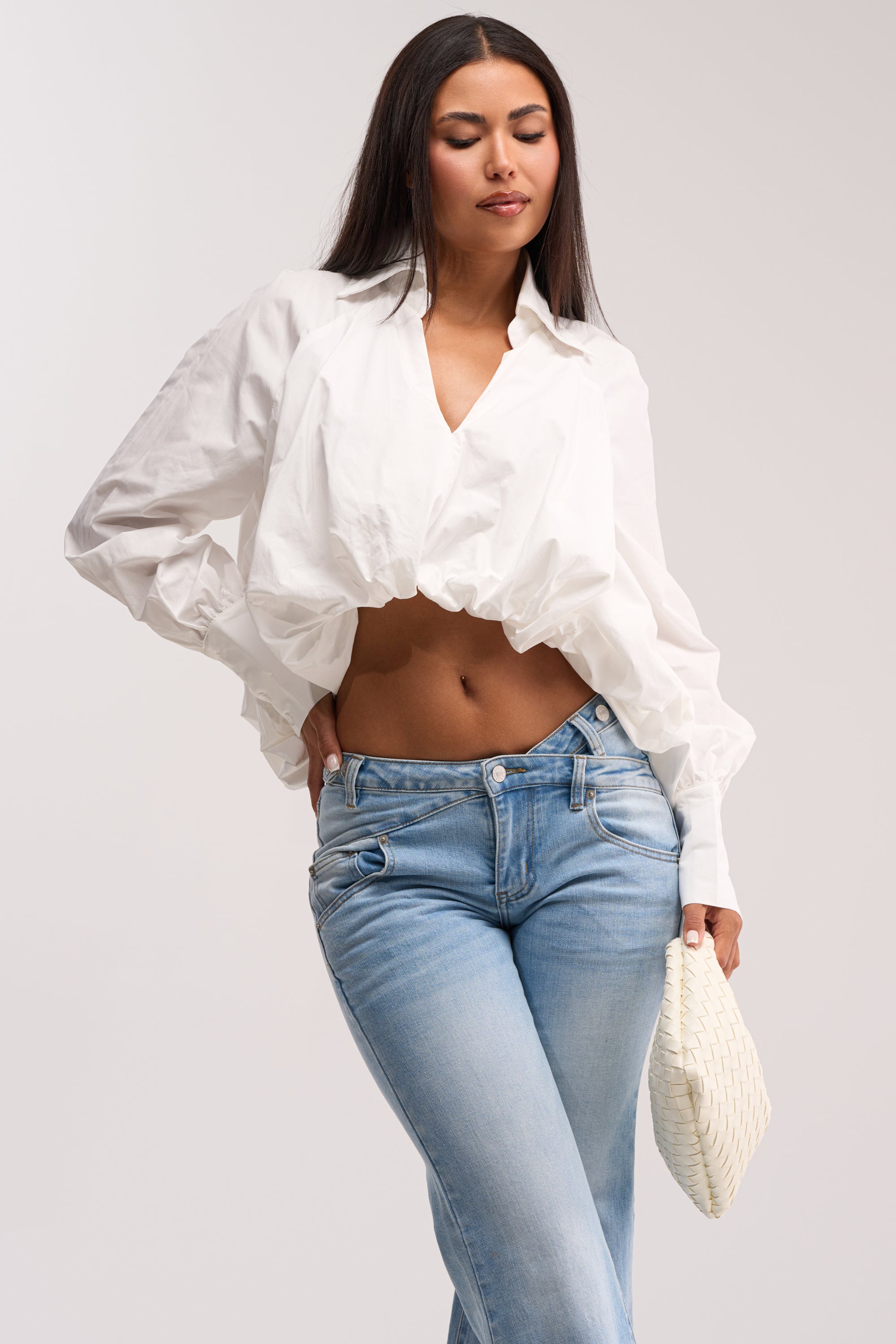 A woman poses in the BEST BEHAVIOR HIGH LOW POPLIN TOP, a white long-sleeve blouse, paired with light blue jeans and holding a small white textured clutch against a plain light background.