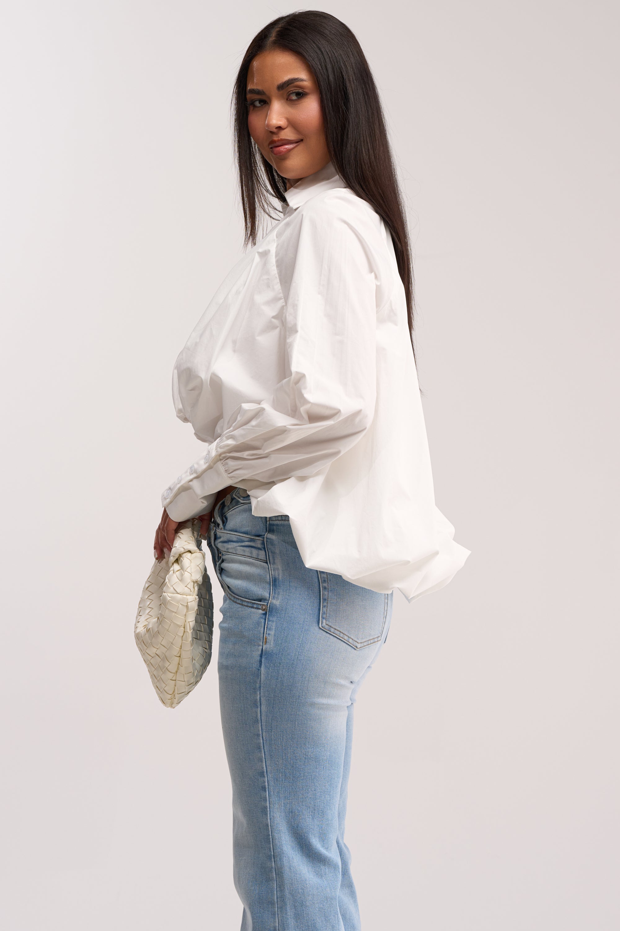 A woman with long dark hair wears the BEST BEHAVIOR HIGH LOW POPLIN TOP in white and light blue jeans, holding a textured white clutch. She stands in profile against a plain light background, looking toward the camera with a slight smile.