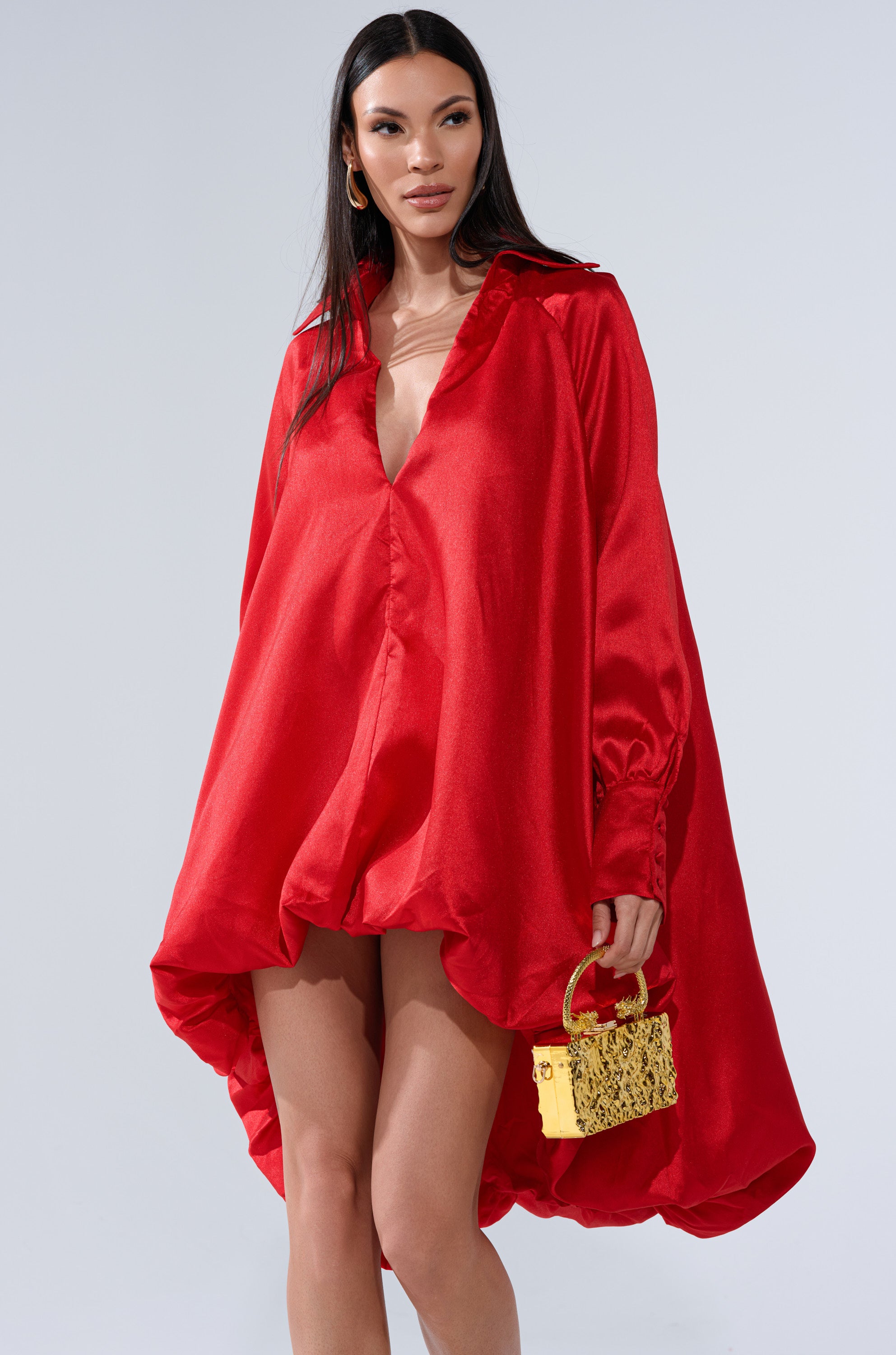Wearing the ELOUISE HI LOW DRESS IN RED, a woman with long dark hair accessorizes with a small yellow textured handbag and gold hoop earrings for an elegant, striking look.