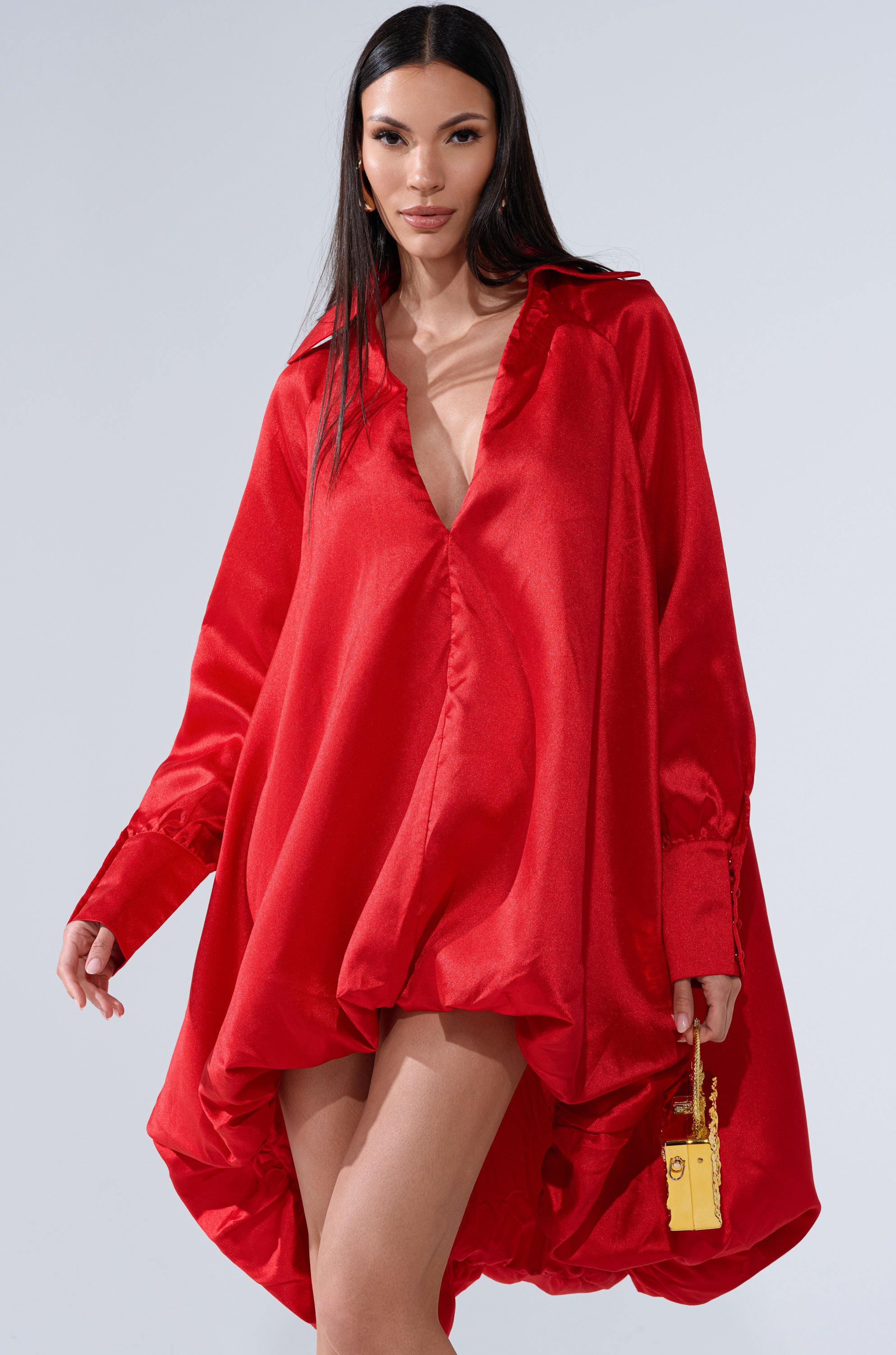 A woman wearing the ELOUISE HI LOW DRESS IN RED—featuring a loose fit, long sleeves, and a deep V neckline—stands against a plain light gray background, holding a small yellow handbag.