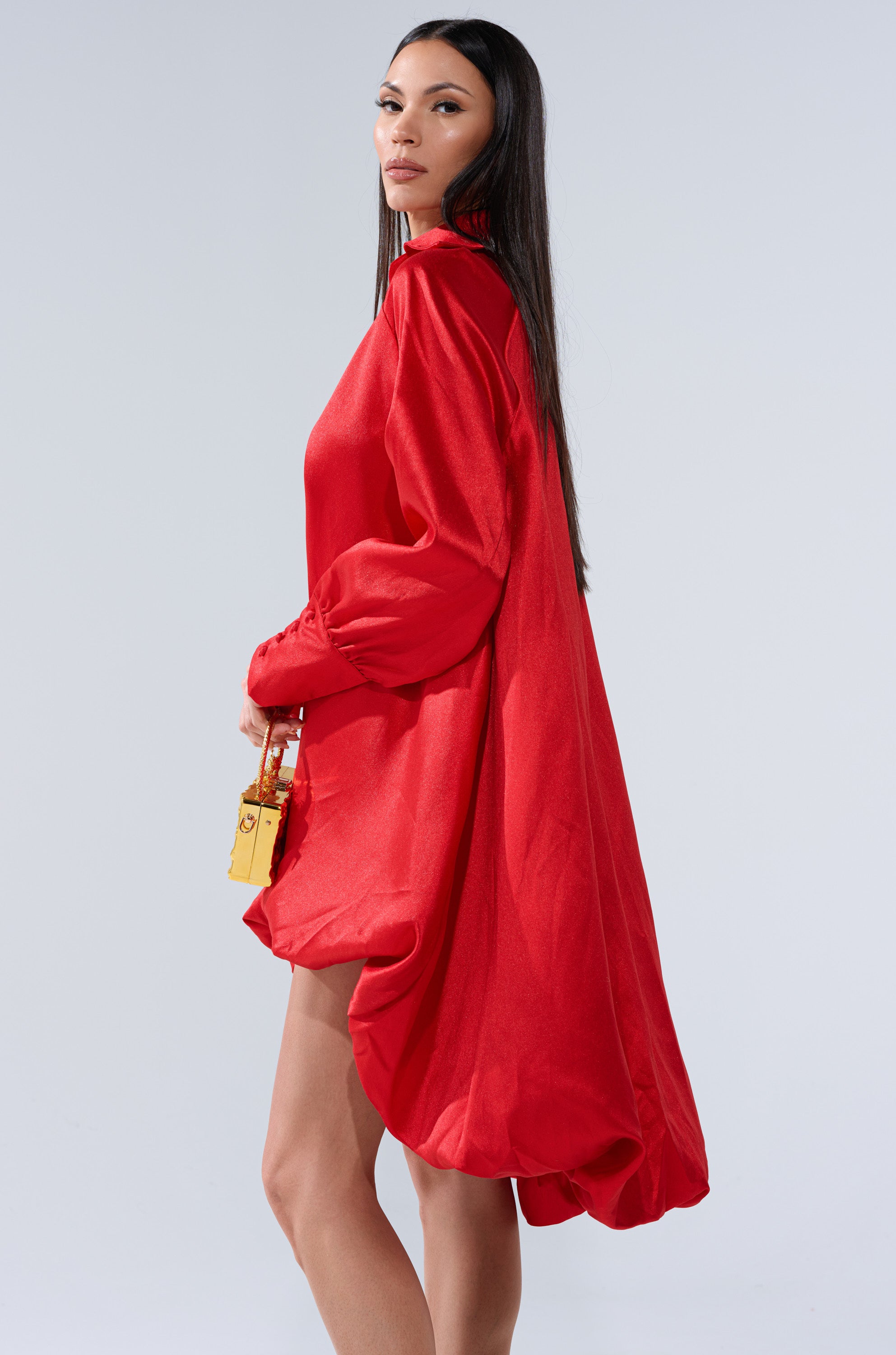 A woman with long dark hair models the ELOUISE HI LOW DRESS IN RED, a loose, flowing evening piece, while holding a small shiny gold handbag against a plain light background.