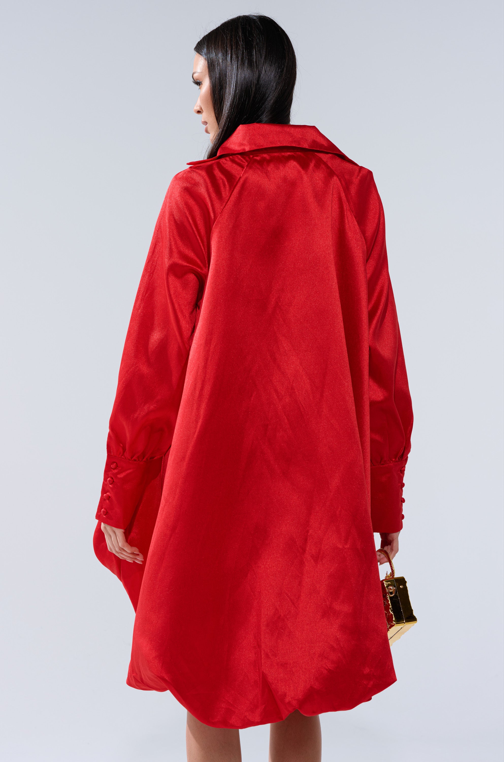 A woman with straight dark hair, seen from behind, wears the ELOUISE HI LOW DRESS IN RED and holds a small gold handbag against a plain light background.