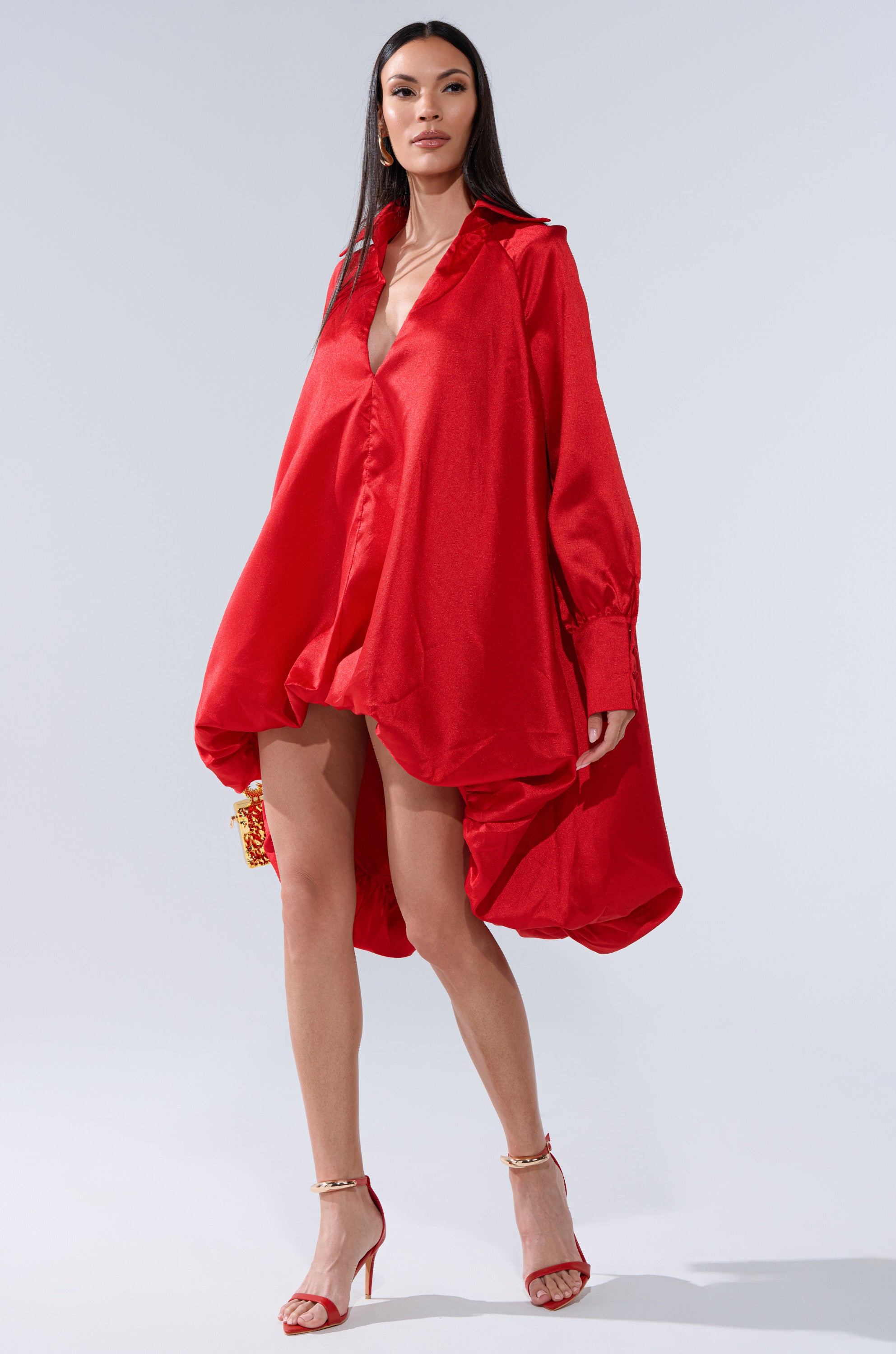 A woman stands confidently in the ELOUISE HI LOW DRESS IN RED, a loose, shiny satin dress with long sleeves, paired with red high-heeled sandals and a small red clutch. The background is plain light grey.