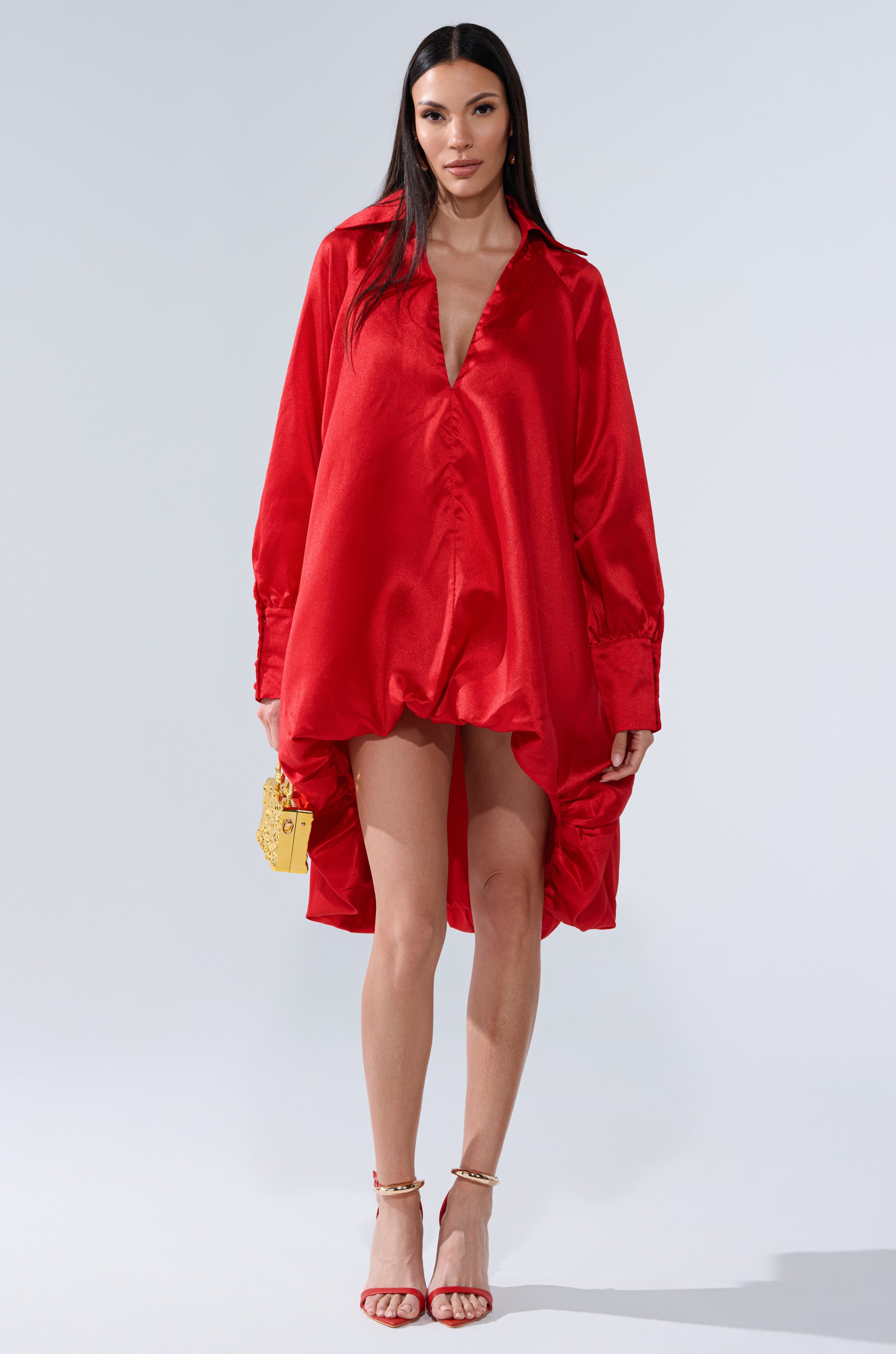 A woman stands against a plain background wearing the ELOUISE HI LOW DRESS IN RED, a loose satin statement dress with a deep V-neck, paired with red high-heeled sandals and a small yellow handbag.