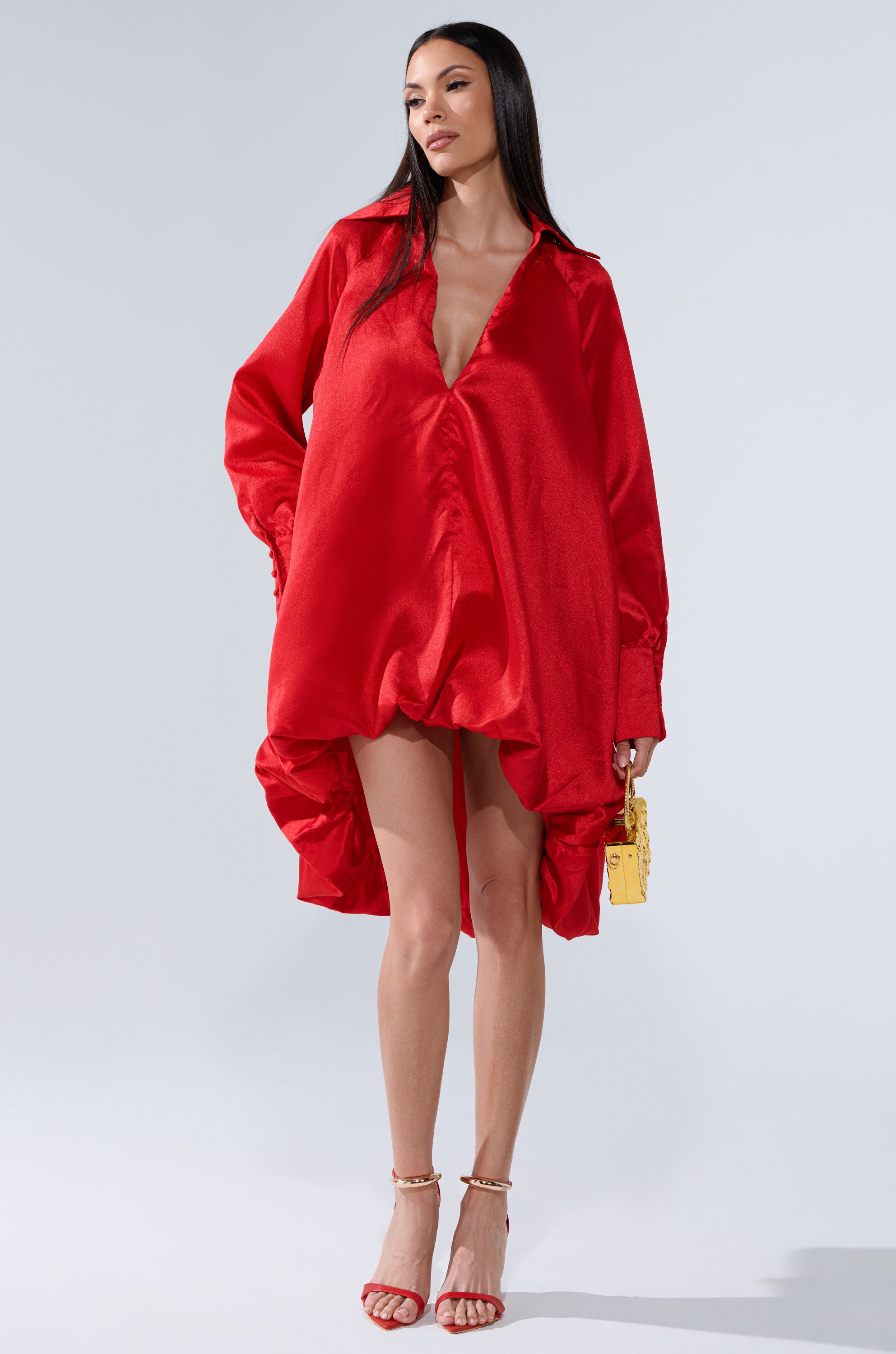 A woman stands against a plain light background wearing the ELOUISE HI LOW DRESS IN RED with a deep V-neckline, paired with strappy red heels and a small yellow handbag.